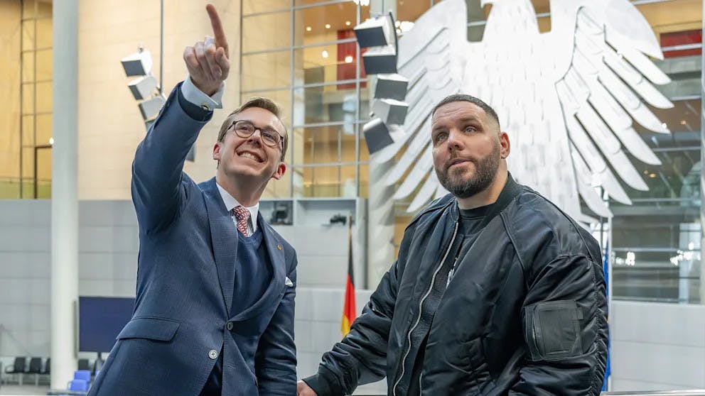 Philipp Amthor zeigt breit grinsend nach oben, rechts neben ihm steht der Rapper Fler. Sie sind im Bundestag