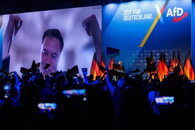 Elon Musk auf einer riesigen Leinwand zu Afd-Veranstaltung zugeschaltet. Er lächelt ausgelassen. Eine Flagge ist direkt unter seiner Nase und sieht aus wie ein Hitlerbart