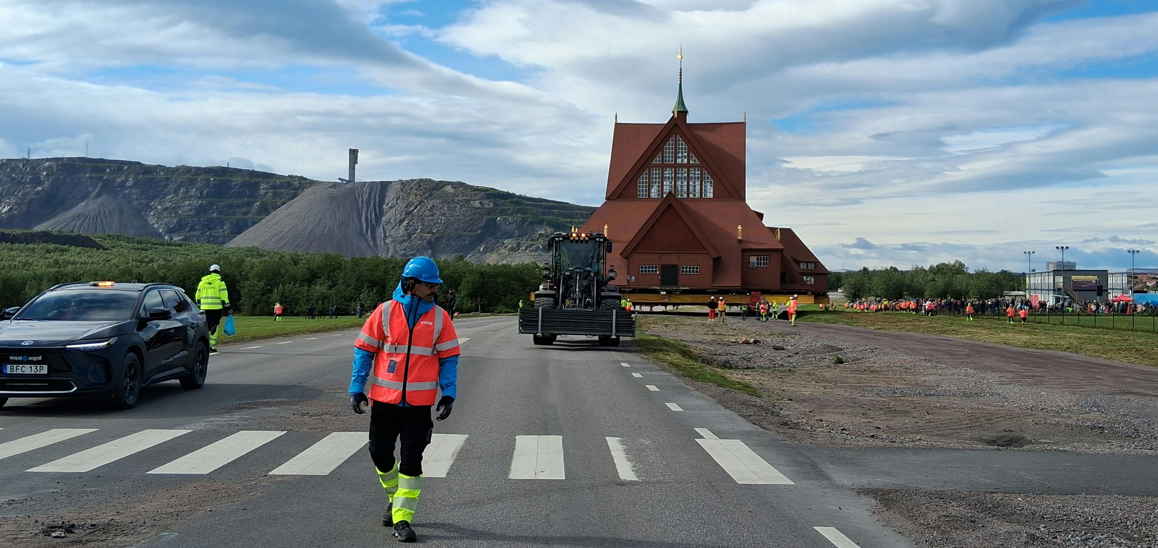 Straße, eine gehende Person in Warnkleidung, dahinter eine Kirche, im Hintergrund ein Berg