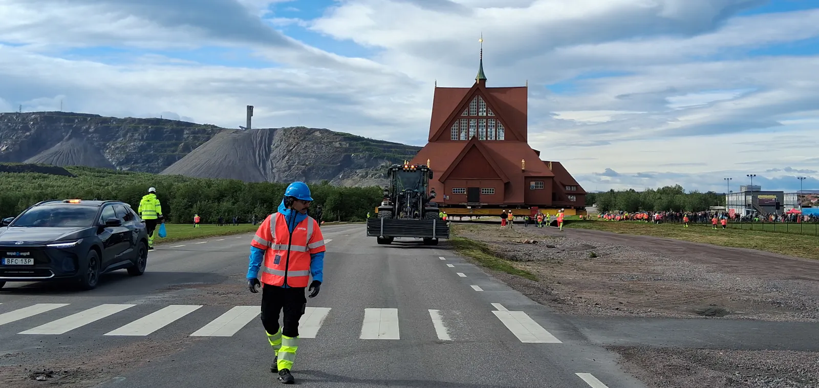 Straße, eine gehende Person in Warnkleidung, dahinter eine Kirche, im Hintergrund ein Berg