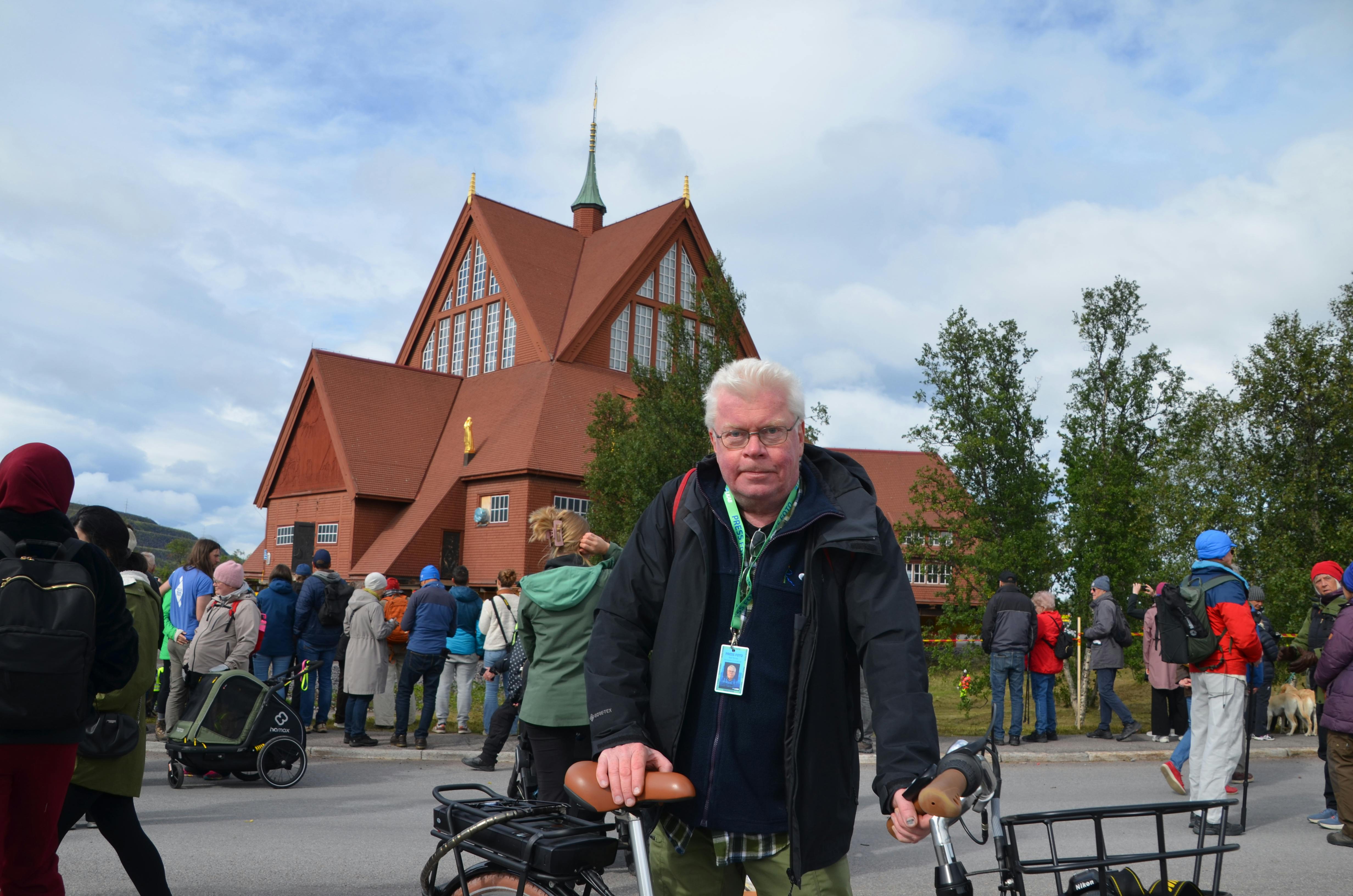 Mann mit Fahrrad vor roter Kirche, viele Leute im Hintergrund