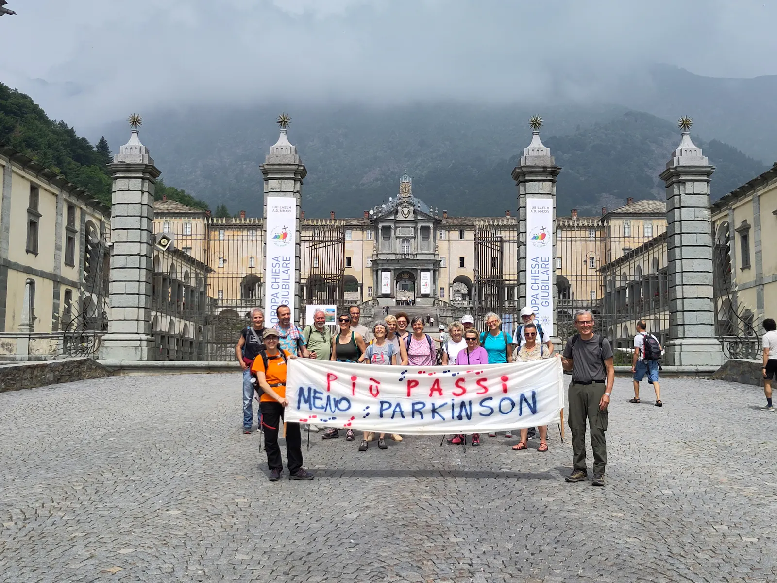 Arrivo di Senza tremori ad Oropa. Striscione retto da giovane a sinistra e non più giovane a destra. Dietro lo striscione il gruppo e sullo sfondo il Santuario di Oropa