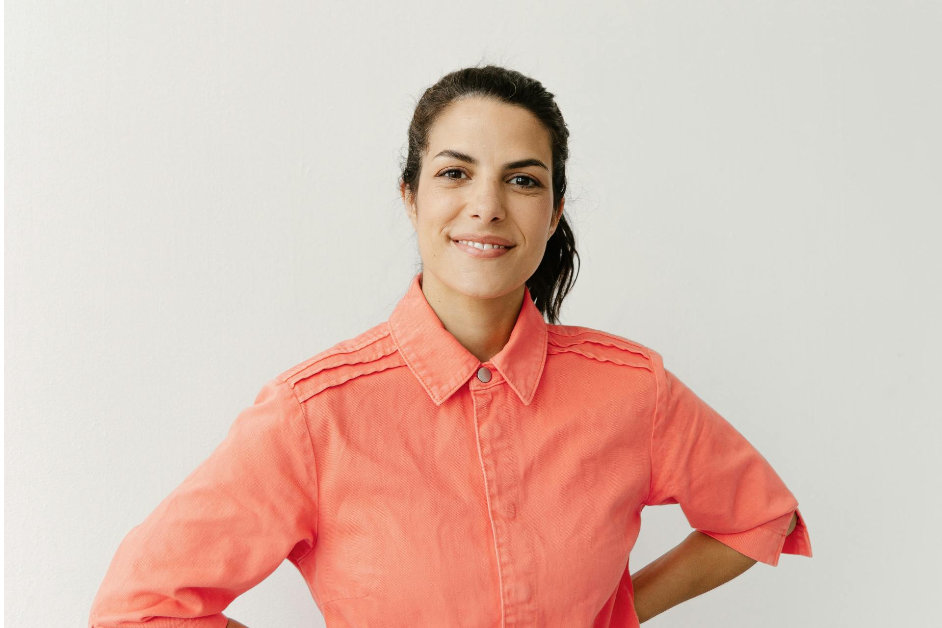 Clarissa Corrêa da Silva trägt ein pastell orange farbenes Hemd, die Hände in die Hüfte gestemmt und lächelt in die Kamera. Die Wand hinter ihr ist weiß.
