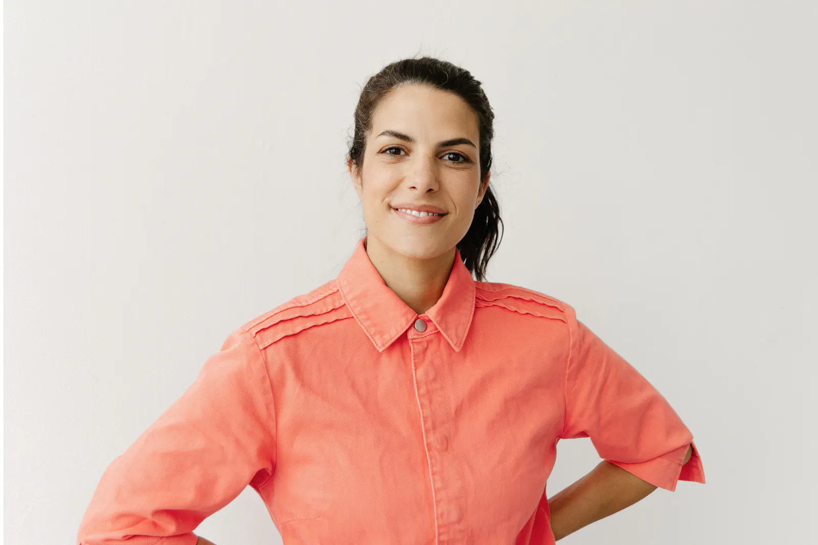 Clarissa Corrêa da Silva trägt ein pastell orange farbenes Hemd, die Hände in die Hüfte gestemmt und lächelt in die Kamera. Die Wand hinter ihr ist weiß.