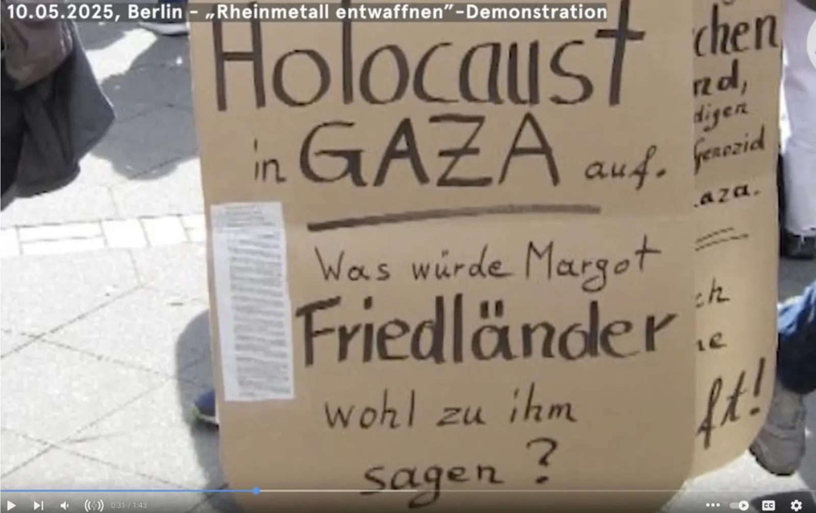 Demonstrationsplakat bei der „Rheinmetall entwaffnen“-Kundgebung am 10. Mai 2025 in Berlin. Auf dem braunen Pappschild steht handgeschrieben: „Holocaust in Gaza auf... Was würde Margot Friedländer wohl zu ihm sagen?“ Der Holocaust wird dabei mit dem Krieg in Gaza gleichgesetzt, Friedländers Name hervorgehoben. Menschen laufen im Hintergrund über das Pflaster, ein Teil eines zweiten Plakats ist am Rand sichtbar.