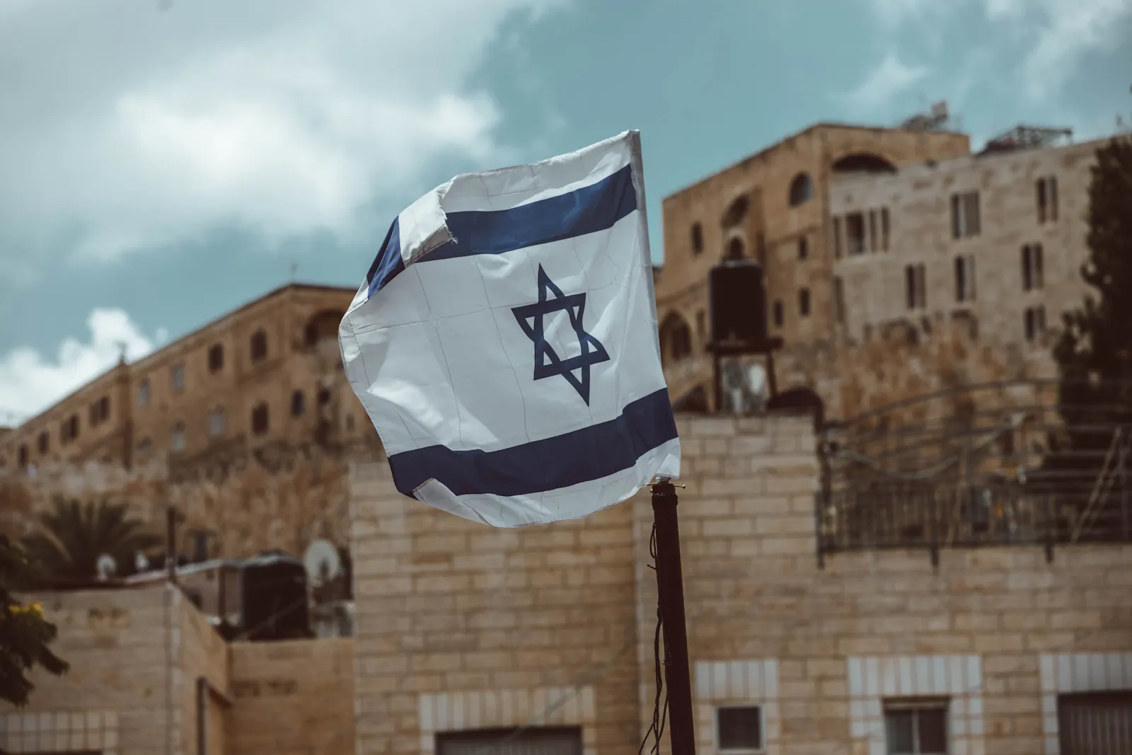 Die israelische Flagge flattert vor einem sandsteinfarbenen Gebäude bei blaugrauem Himmel.