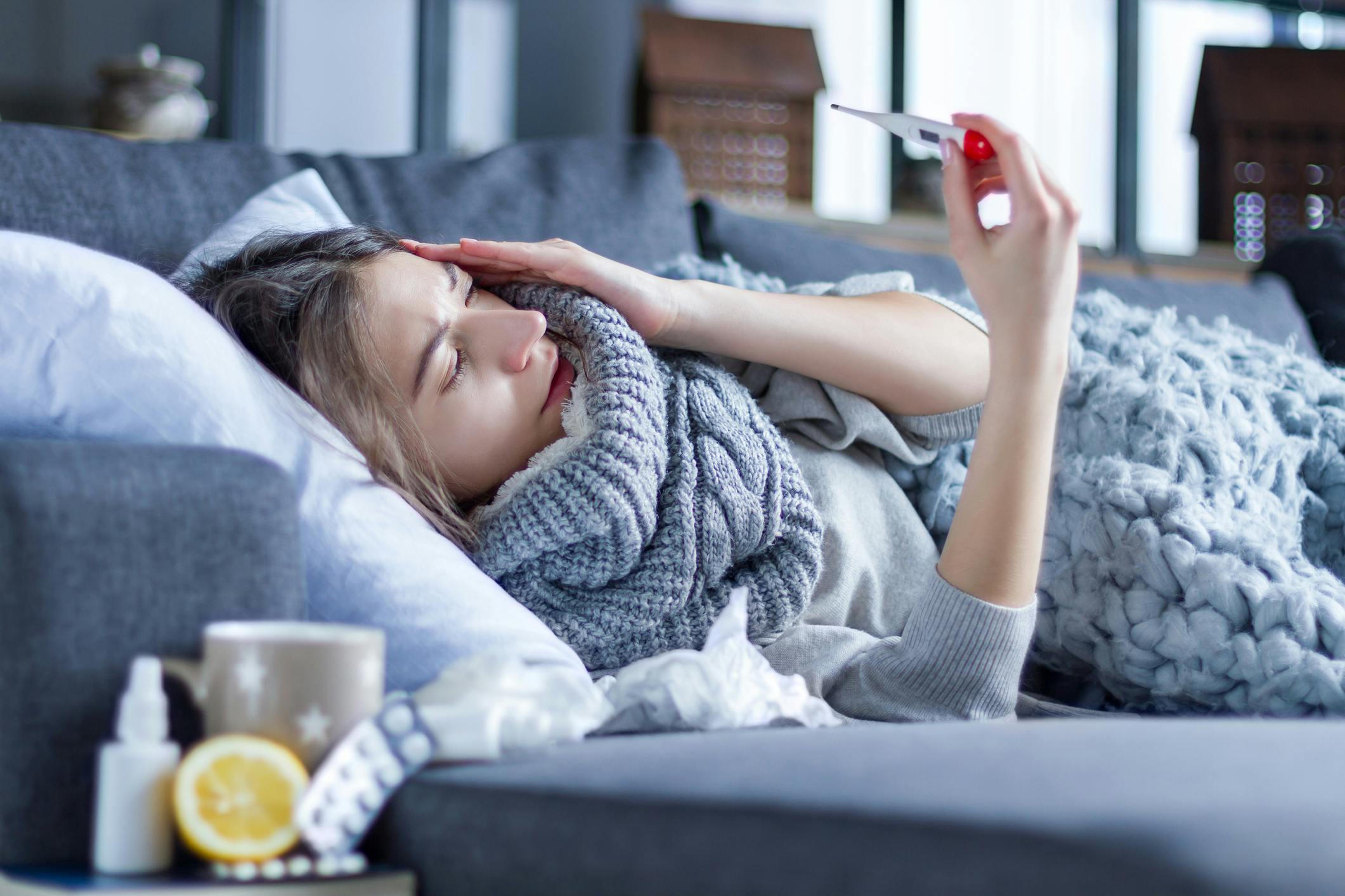 Das Foto zeigt eine kranke Frau, die im Bett liegt. Sie hält ein Fieberthermometer in der Hand. Neben ihr stehen Medikamente, eine Tasse Tee und eine Zitrone.