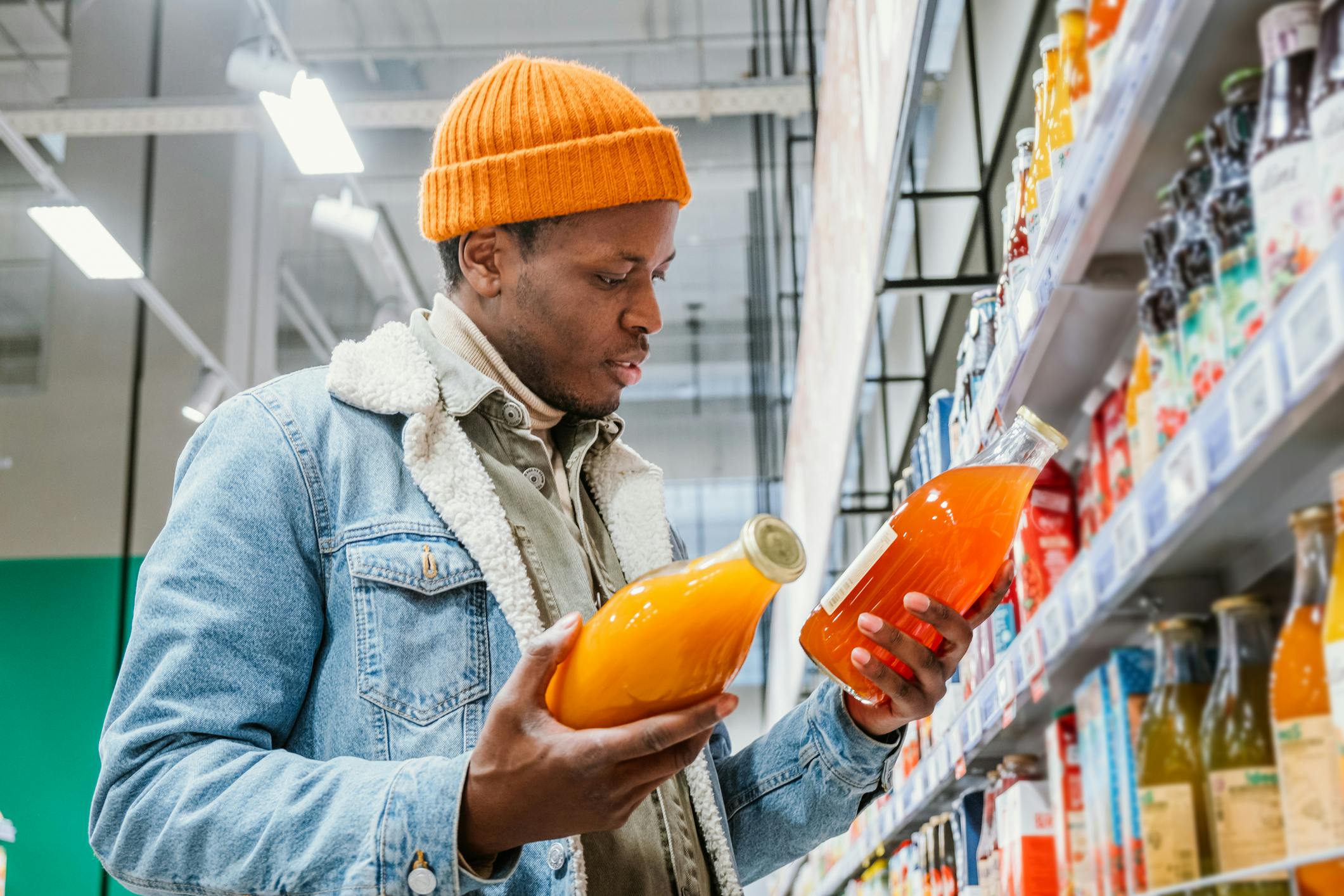 Dieses Foto zeigt einen jungen Mann im Supermarkt, der zwei orangefarbene Saftflaschen vergleicht.