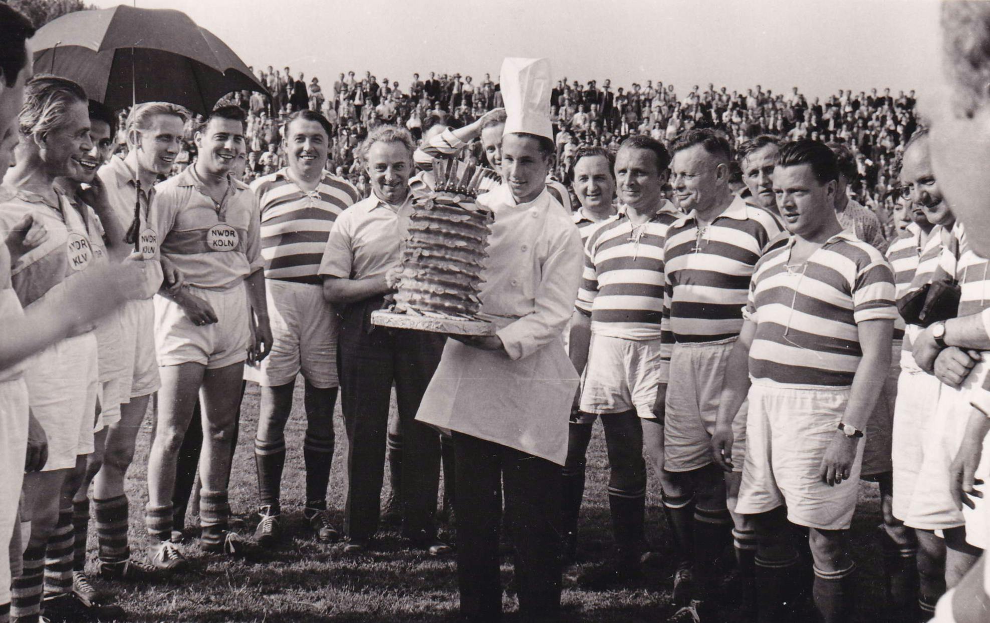 Schwarz-weiß Foto mit Fußballspielern auf dem Rasen. In der Mitte ein Konditor mit weißer Mütze, der einen riesigen Baumkuchen mit Krone trägt.
