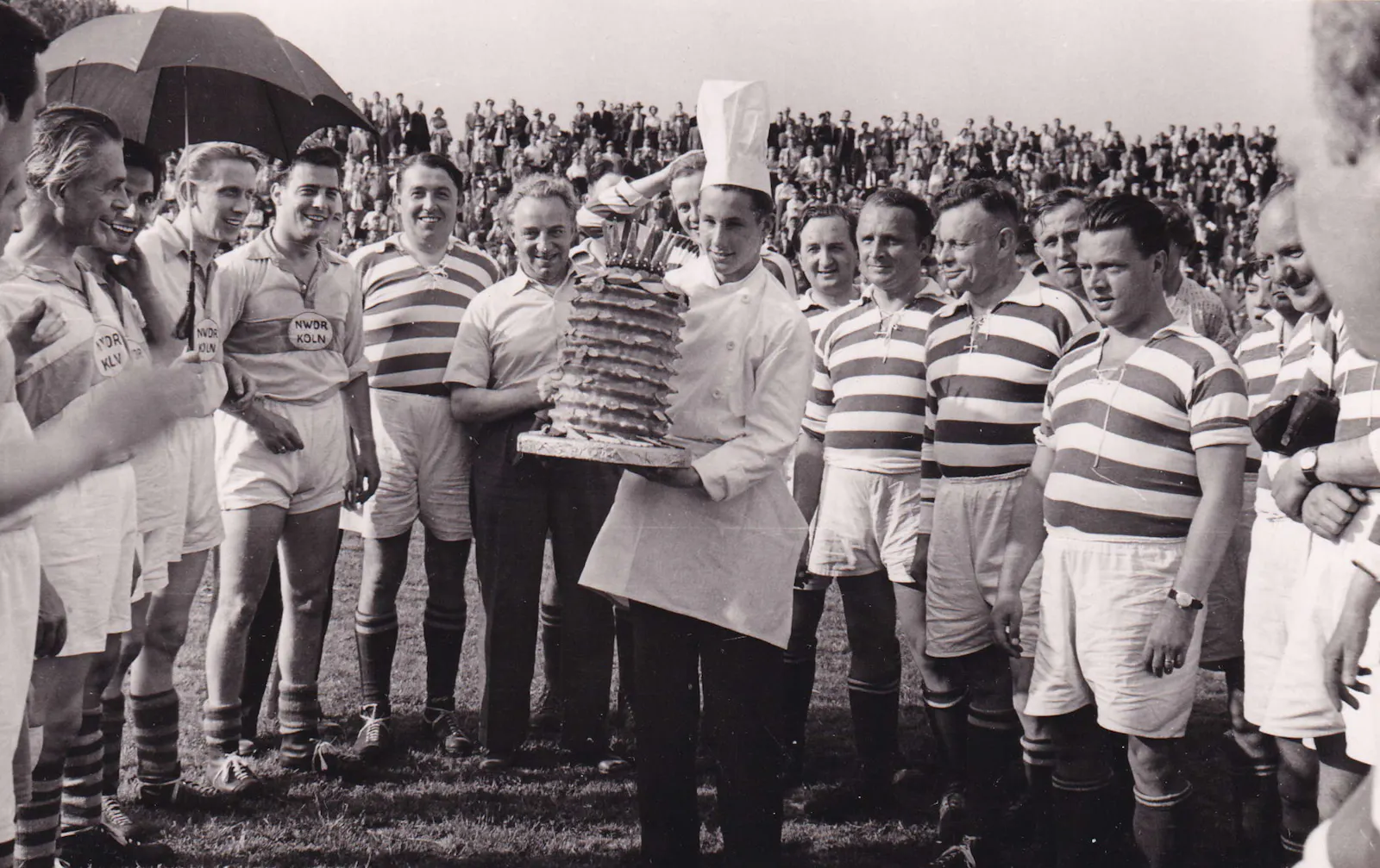 Schwarz-weiß Foto mit Fußballspielern auf dem Rasen. In der Mitte ein Konditor mit weißer Mütze, der einen riesigen Baumkuchen mit Krone trägt.