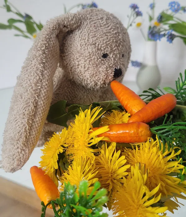 Stoffhase Benny mümmelt Plastikmöhrchen und gelben Löwenzahn.