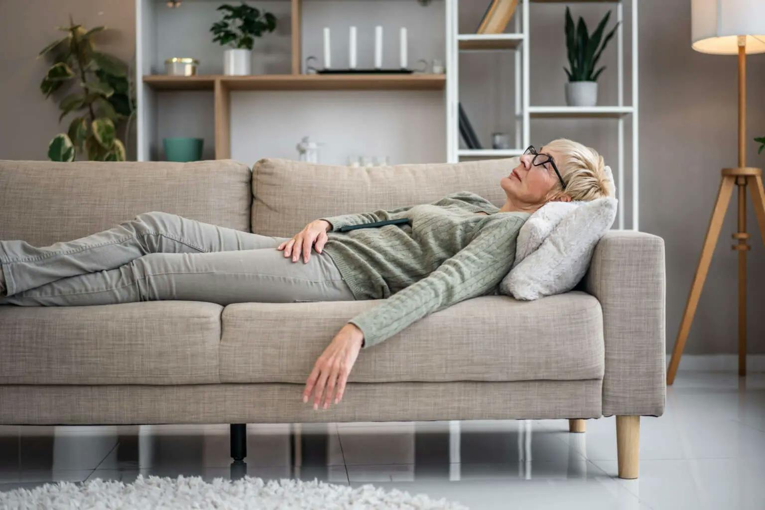Ältere Frau mit Kurzhaarschnitt und Brille liegt erschöpft auf dem Sofa