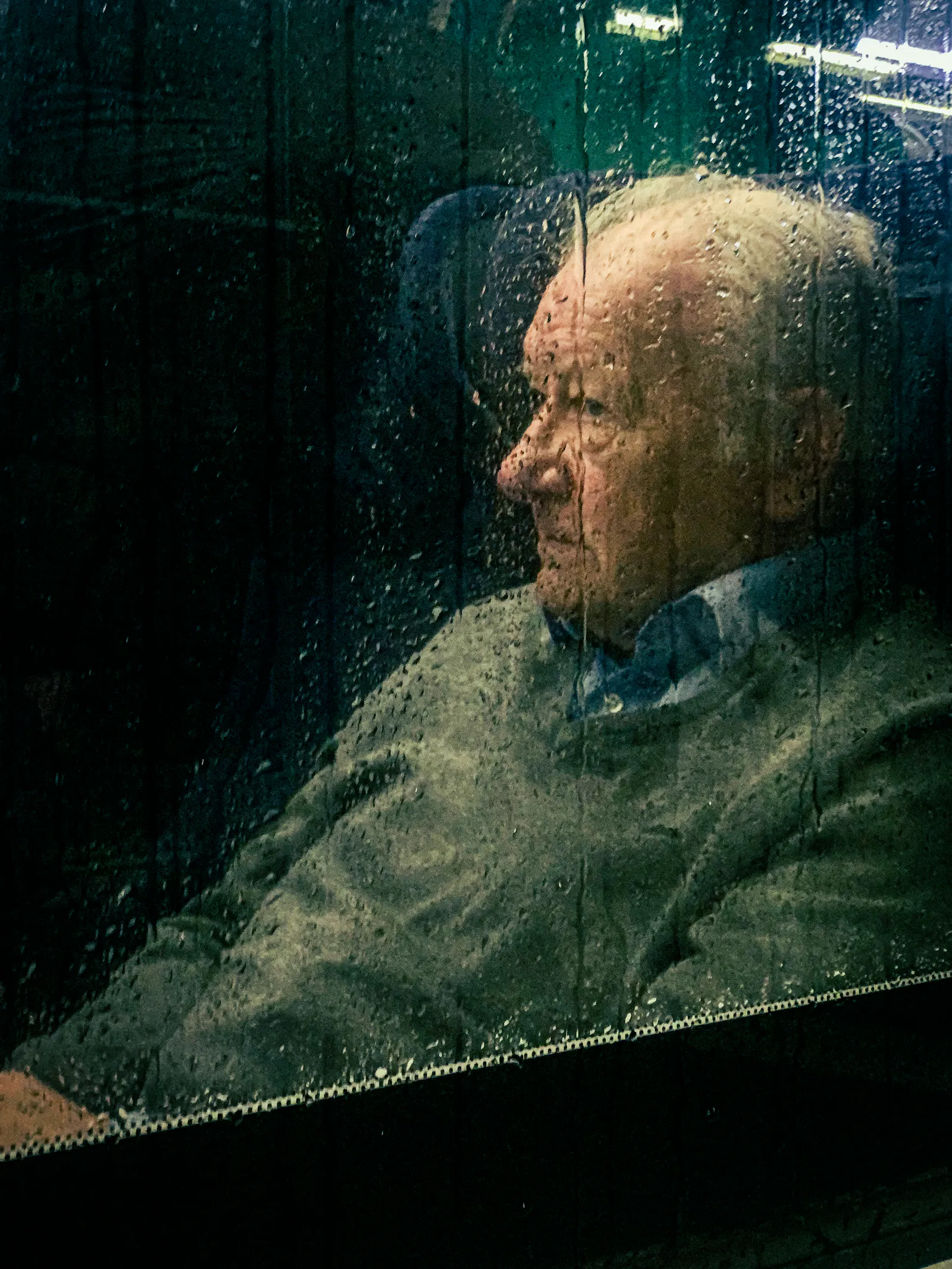 Ein älterer Herr sitzt hinter einer verregneten Glasscheibe und schaut etwas grimmig.