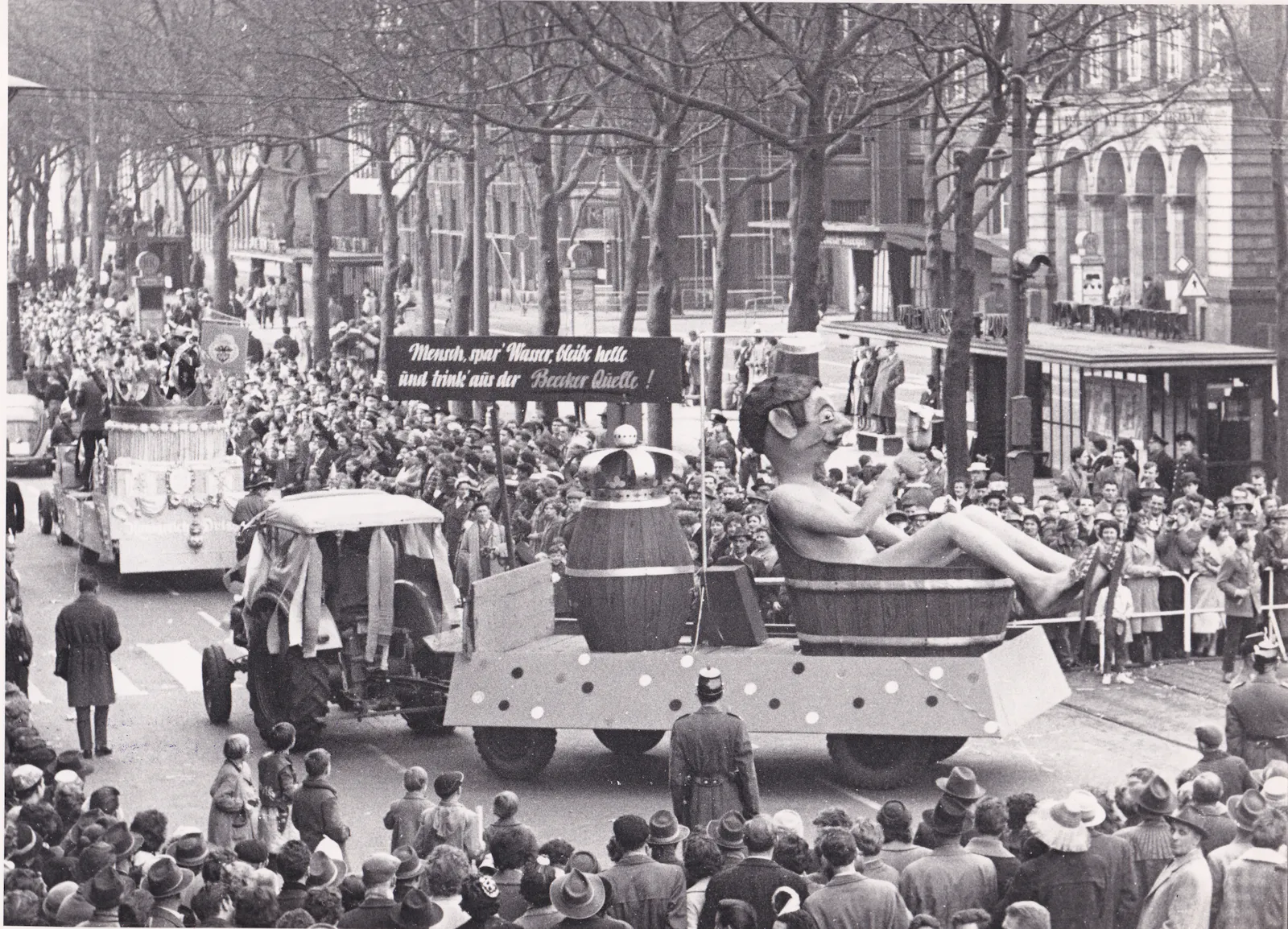 Schwarz-Weißfoto eines Karnevalsumzuges in der Duisburger Innenstadt aus den 1950er Jahren. Zu sehen der Prunkwagen der König-Brauerei. mit einem Fass Bier und einer männlichen Puppe, die in einem Holzschuber sitzt und badet. Darüber steht der Spruchband: "Mensch, spar Wasser, bleibe helle und trink aus der Beecker Quelle"