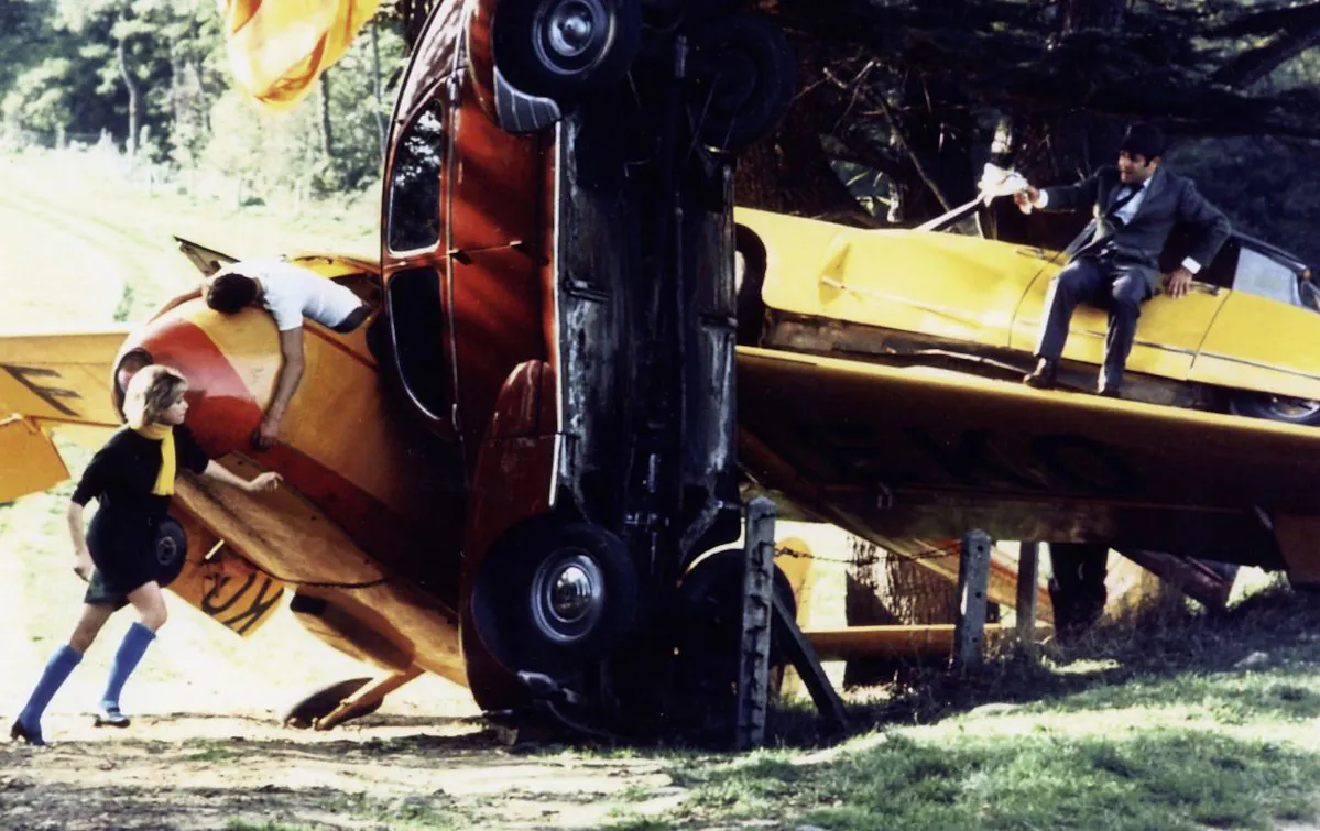 Filmstill: Eine groteske Massenkarambolage, Autos, die sich stapeln, auf einem sitzt ein Mann, von links läuft eine Frau auf ihn zu.