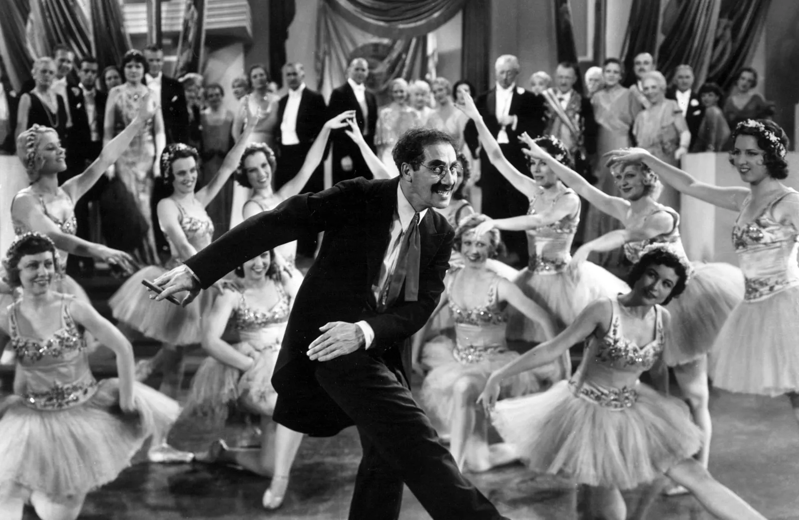 Filmstill aus "Duck Soup": Groucho Marx im Frack verrenkt sich im Tanz, umgeben von Balletteusen, hinten Gäste eine schicken Gesellschaft.