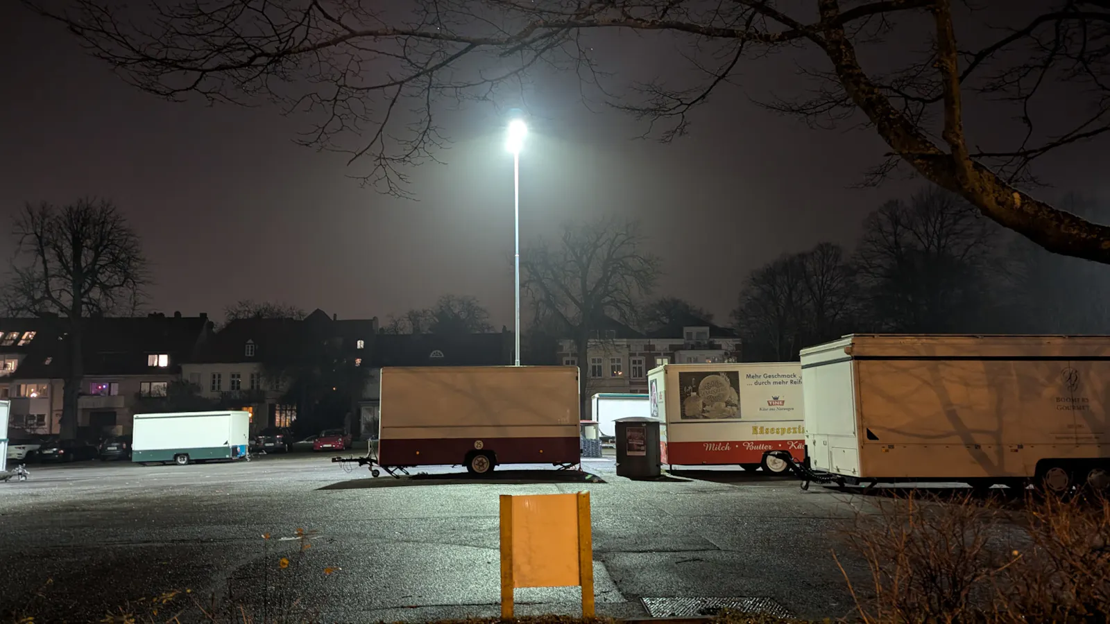 Ein geschlossener Wochenmarkt in einer deutschen Stadt am Abend. Verrammelte Marktwagen im Flutlicht einer Laterne, dahinter Häuser, kahle Bäume.
