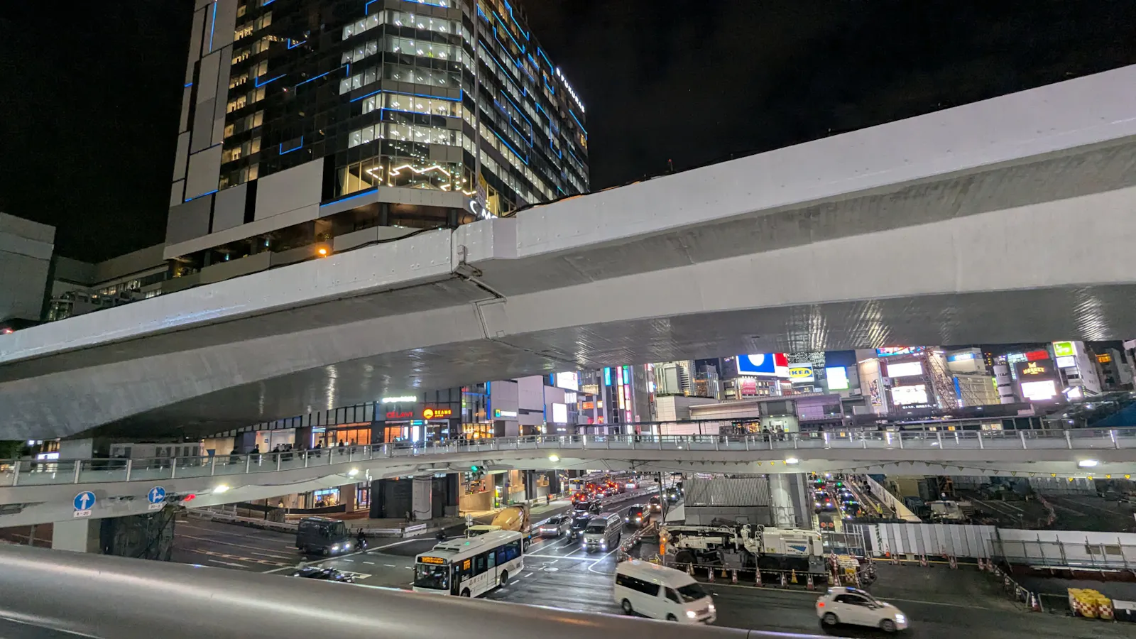 Shibuya, Tokio, am Abend. Eine Hochstraße, eine Fußgängerüberführung, eine Straße, dahinter Hochhäuser mit LED-Screens an den Fassaden, Lichtgewitter.