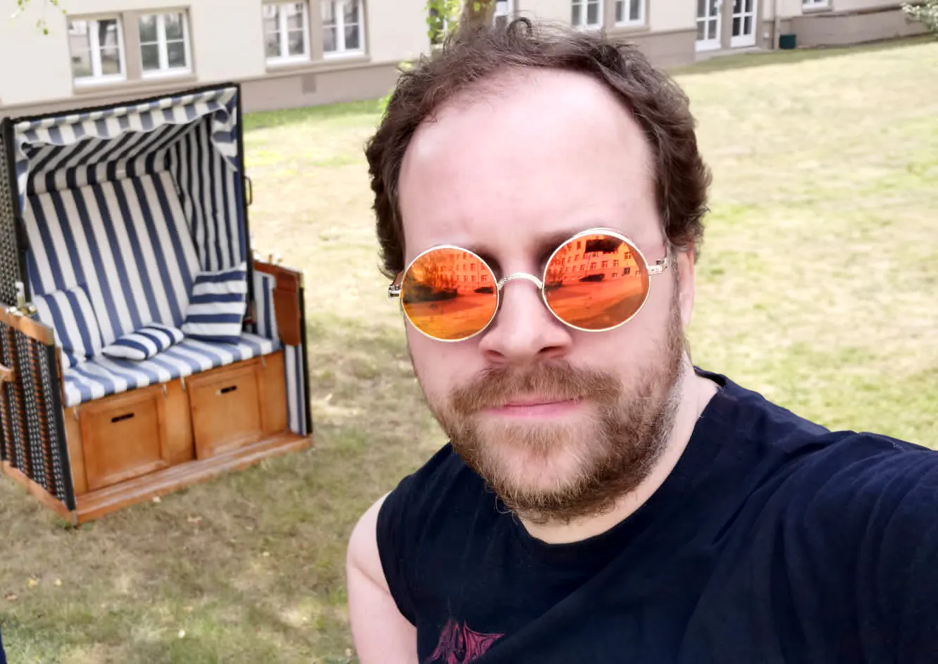 Unser Professor Doktor Spielleiter, der mit cooler orangener Sonnenbrille vor einem Strandkorb steht.