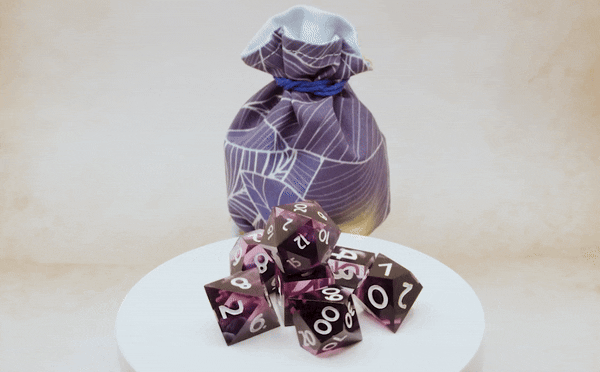 Auf einem Drehteller sieht man die wunderschönen lilaglänzenden Würfel von Cat in the Dice Bag und dahinter steht ein passender lilafarbener Beutel von Fateful Fabrics.