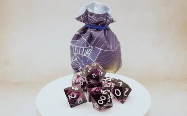Auf einem Drehteller sieht man die wunderschönen lilaglänzenden Würfel von Cat in the Dice Bag und dahinter steht ein passender lilafarbener Beutel von Fateful Fabrics.