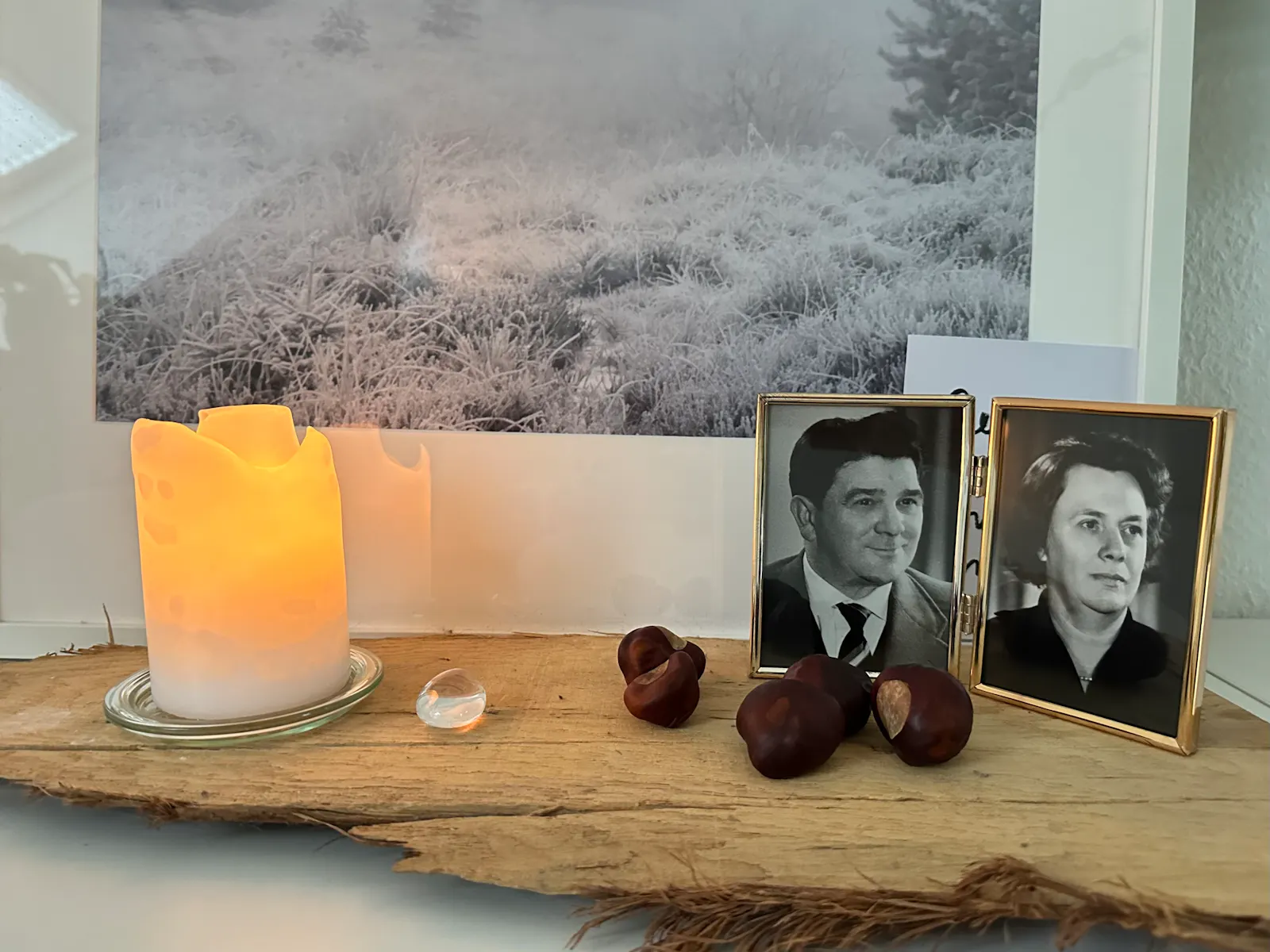 Holzbrett. Darauf stehen eine brennende weiße Stumpenkerze und ein Doppelrahmen mit Schwarz-Weiß-Fotos eines Mannes und einer Frau. Dazwischen liegen ein Stein und Kastanien.