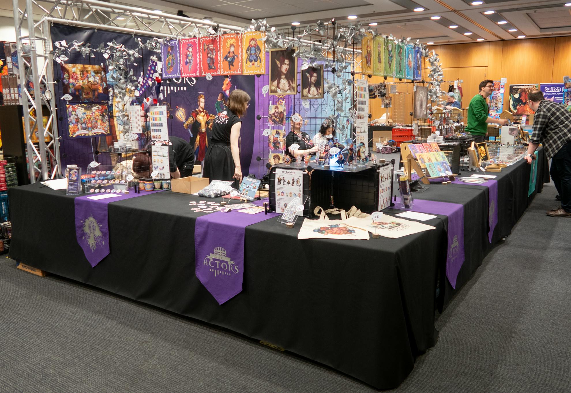 Das Bild zeigt unseren Merch-Stand in einem großen, hell beleuchteten Raum. Der Stand ist mit schwarzen Tischtüchern bedeckt, die mit lila Stoffläufern verziert sind, auf denen das Logo „Dice Actors“ und ein Silberbaum zu sehen ist. Auf den Tischen sind viele kleine Figuren, Kunstwerke und weitere Merchandise-Artikel wie Buttons und Taschen ausgestellt.

Im Hintergrund hängen bunte Bilder von den Hütern der Obelisken an Gittern. Hinter dem Tisch stehen einige unserer Merch-Mausis.