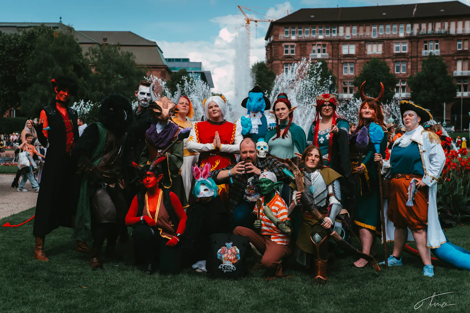 Das Foto zeigt eine Gruppe von etwa 17 Diceys, die sich als Dice Actors Charaktere verkleidet haben. Sie stehen und knien auf einer Wiese vor einem großen Springbrunnen.
Viele tragen Hörner, Elfenohren, Masken oder bunte Gesichter.
Alle wirken fröhlich und posieren für ein Gruppenfoto im Freien. Im Hintergrund sind Blumen, Bäume, alte Gebäude und viele andere Menschen zu sehen.