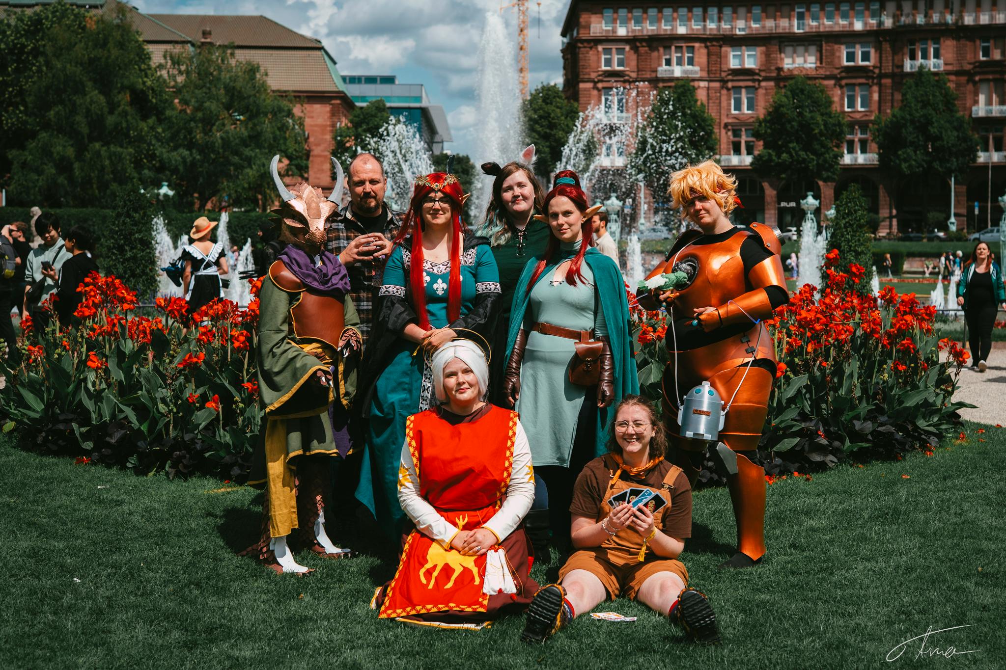 Das Foto zeigt ebenfalls eine Gruppe von neun Diceys, die sich als Jeri, Alex, Elaynie, Daucus und andere Charakter verkleidet haben. Sie stehen oder sitzen auf einer Wiese mit roten Blumen im Hintergrund. Dahinter sind Springbrunnen, Bäume und ein großes, altes Gebäude zu sehen.