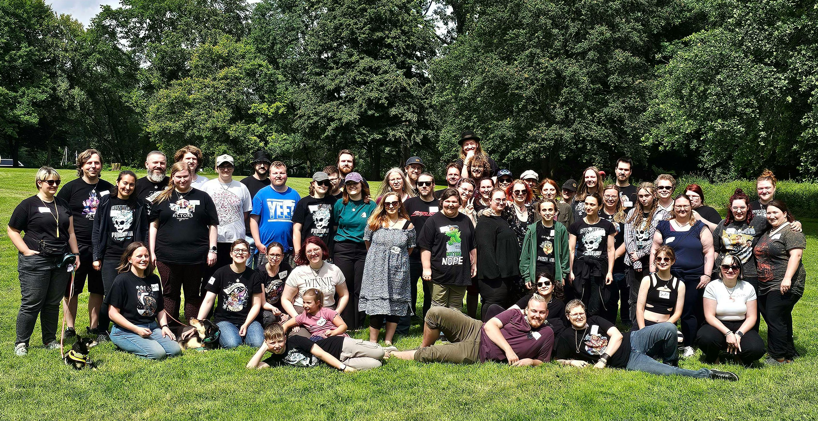 Das offizielle Gruppenfoto des Dice-Actors-Sommertreffens 2026: Eine große Gruppe von über 50 strahlenden Diceys versammelt sich auf einer grünen Wiese unter bewölktem Sommerhimmel. Viele tragen Dice-Actors-Merchandise. Einige sitzen oder liegen auf dem Gras, andere stehen. Hunde sind auch mit dabei. Die Stimmung wirkt ausgelassen und herzlich.