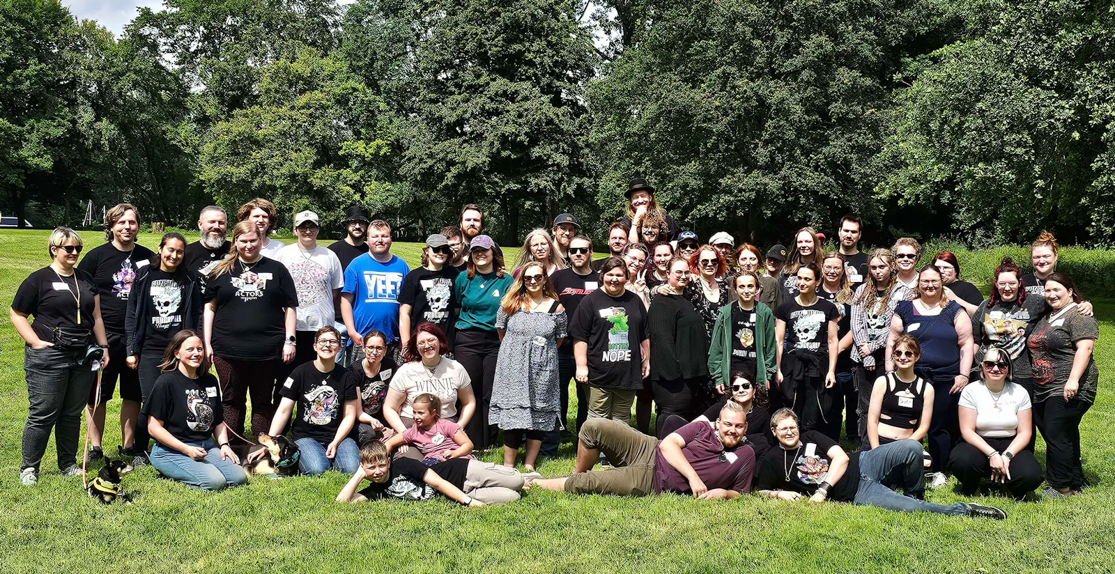 Das offizielle Gruppenfoto des Dice-Actors-Sommertreffens 2026: Eine groĂe Gruppe von ĂŒber 50 strahlenden Diceys versammelt sich auf einer grĂŒnen Wiese unter bewölktem Sommerhimmel. Viele tragen Dice-Actors-Merchandise. Einige sitzen oder liegen auf dem Gras, andere stehen. Hunde sind auch mit dabei. Die Stimmung wirkt ausgelassen und herzlich.