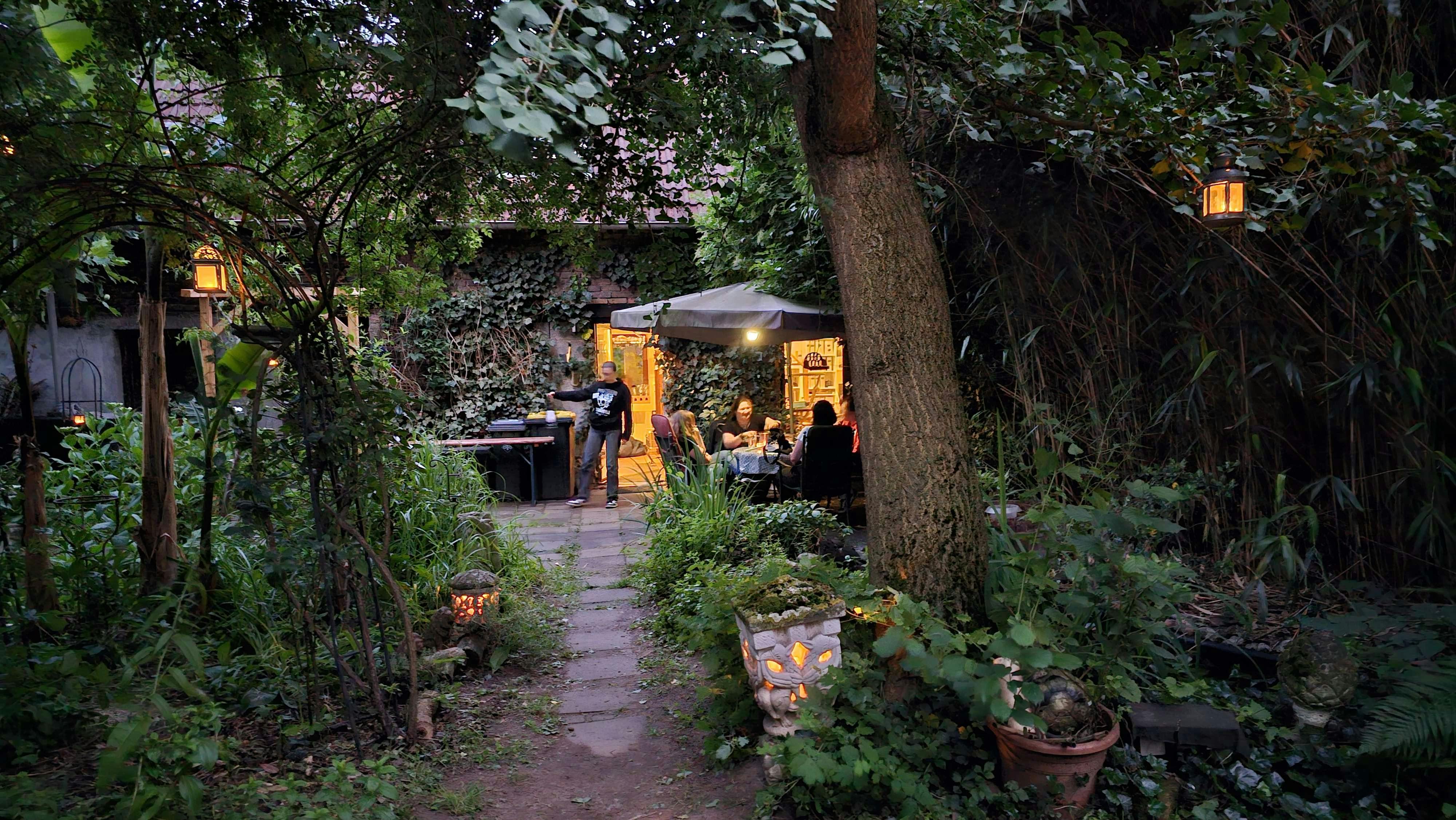 Ein stimmungsvolles Abendfoto von Judiths üppig begrüntem Garten in der Dämmerung. Ein Steinplattenweg führt durch dichte Vegetation aus Efeu und Sträuchern in Richtung einer beleuchteten Terrasse, wo mehrere Personen an einem Tisch unter einem Sonnenschirm sitzen. Entlang des Weges leuchten kleine Laternen und dekorative Steinfiguren mit Kerzen. Im Hintergrund ist ein efeubedecktes Haus zu sehen.