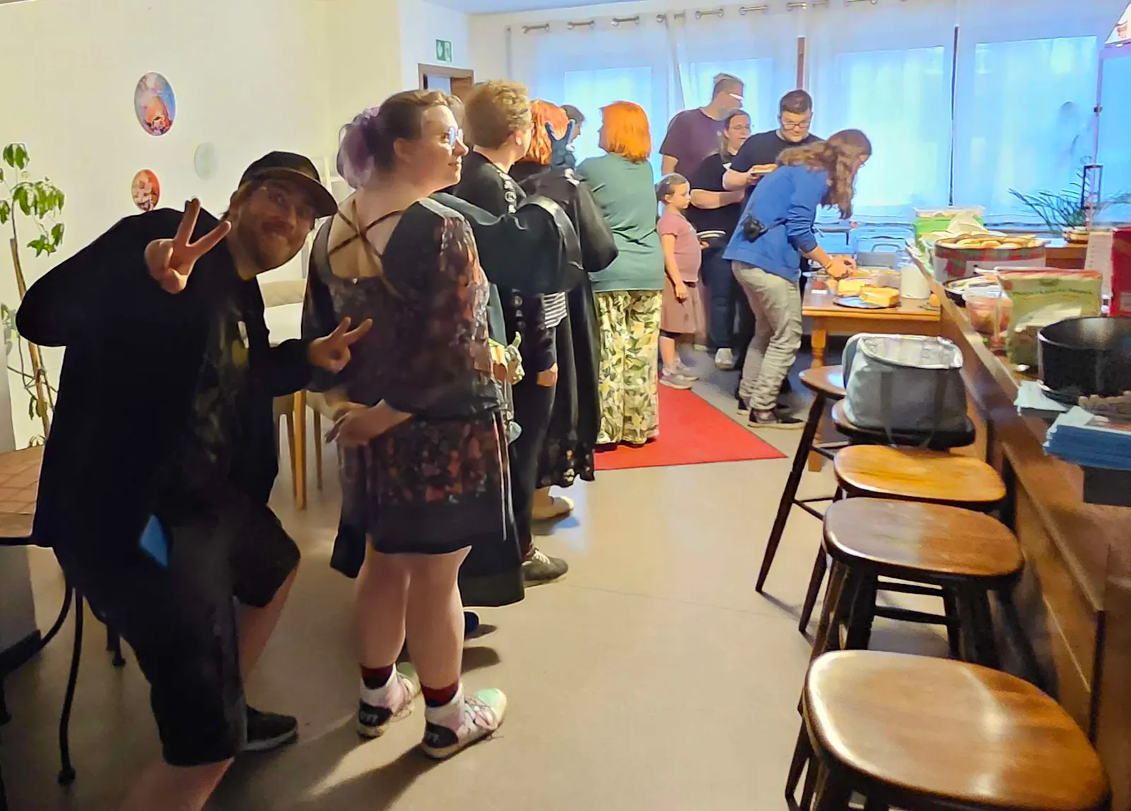 Eine lange Schlange von Menschen steht in einem hellen Raum und wartet auf das Kuchenbuffet, das rechts im Hintergrund aufgebaut ist. Links vorne posiert jemand frech in die Kamera. An der Wand hÀngen bunte Illustrationen, auf dem roten Teppich stehen weitere GÀste. Der Raum ist gut besucht und die Stimmung wirkt ausgelassen und fröhlich.