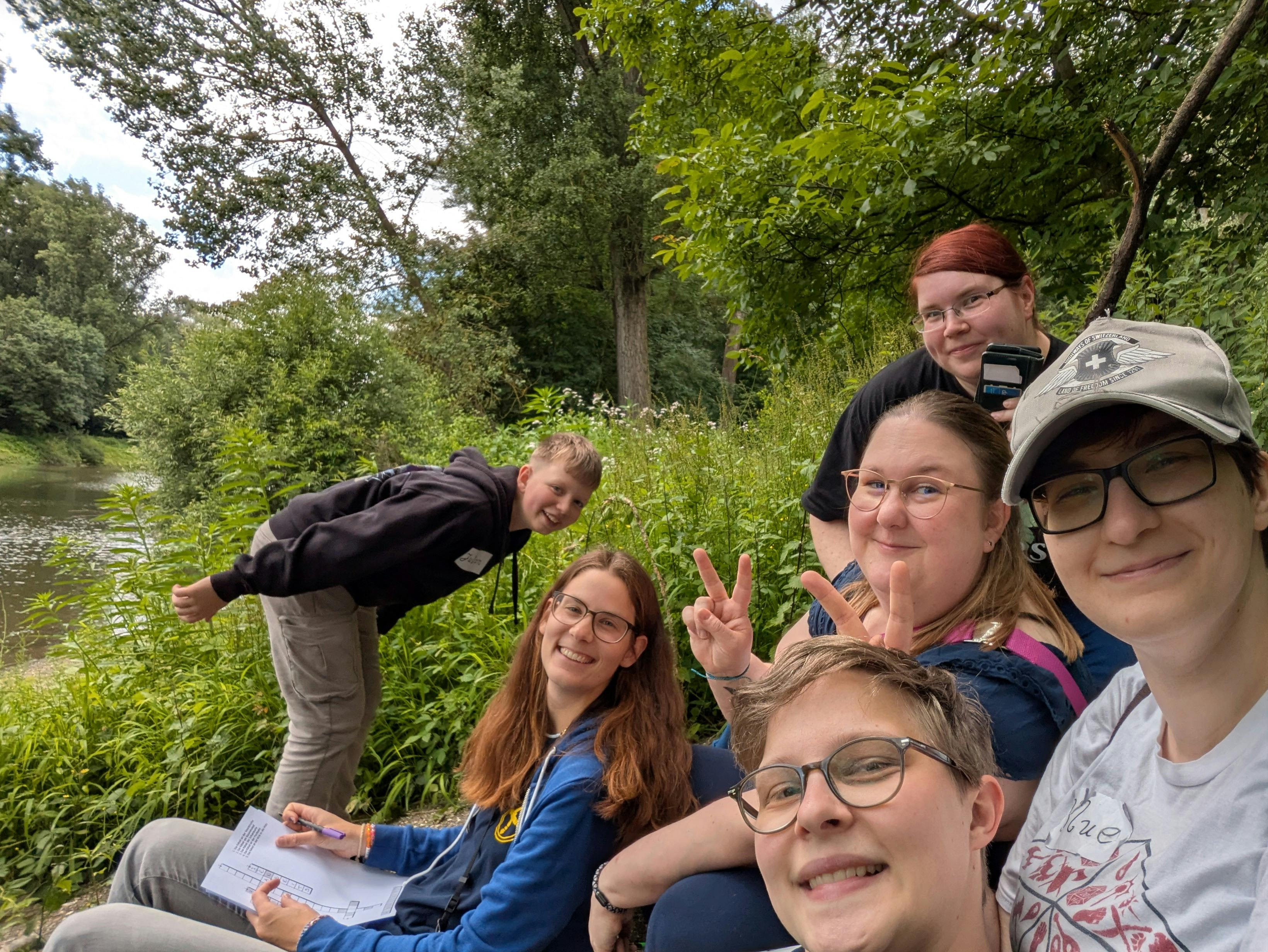 Ein Gruppenselfie von sechs Diceys in der Natur an einem Flussufer, umgeben von üppigem Grün. Links balanciert jemand auf einem Stein und lächelt in die Kamera. Die anderen fünf Personen – alle mit Brille, teils mit Cap – lachen, zeigen Peace-Zeichen und blicken fröhlich in die Kamera. Eine Person hält ein Blatt mit einem Rätsel oder einer Quest-Aufgabe in der Hand. Im Hintergrund fließt ein ruhiger Fluss durch die grüne Landschaft.
