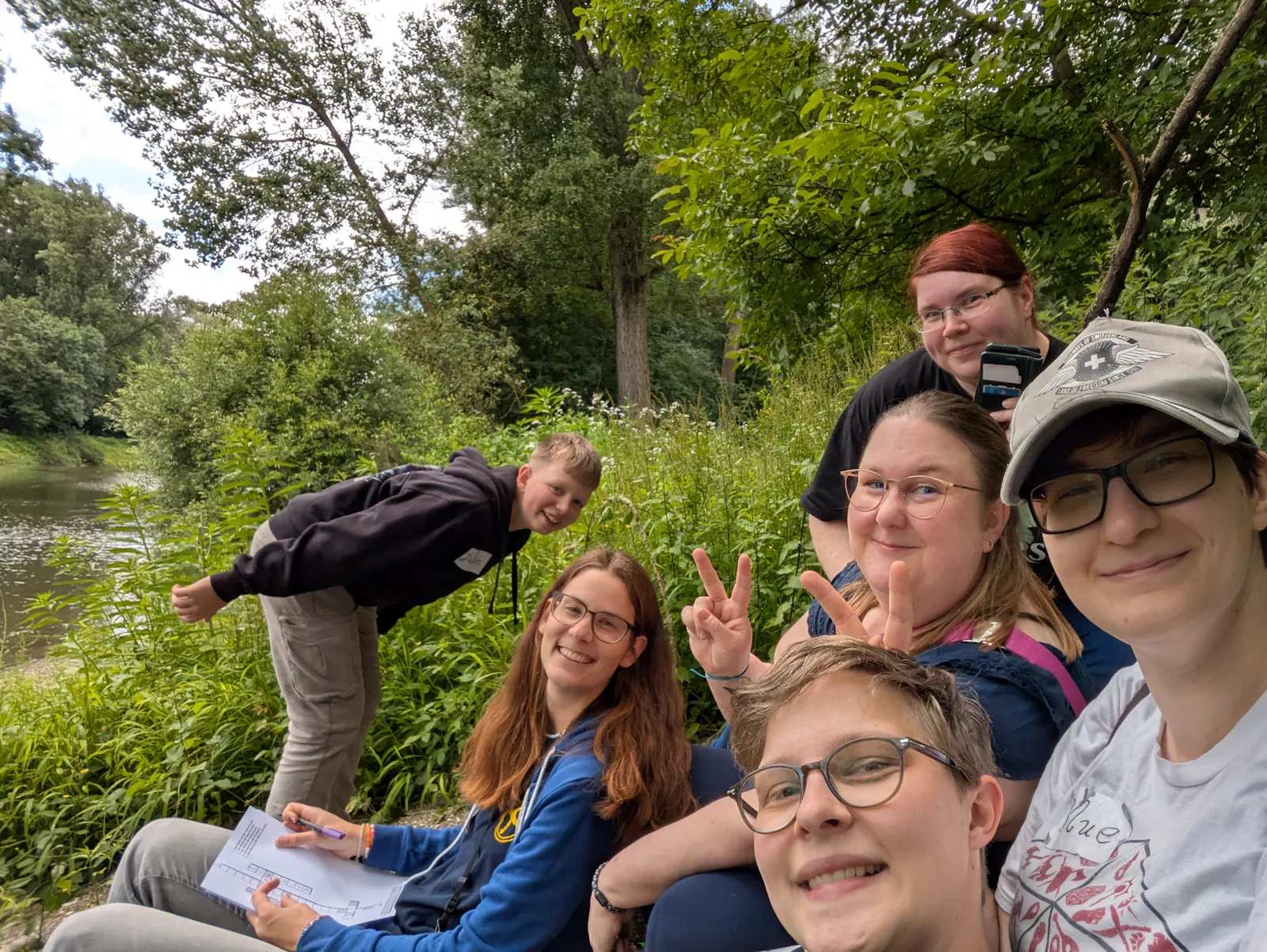 Ein Gruppenselfie von sechs Diceys in der Natur an einem Flussufer, umgeben von ĂŒppigem GrĂŒn. Links balanciert jemand auf einem Stein und lĂ€chelt in die Kamera. Die anderen fĂŒnf Personen â alle mit Brille, teils mit Cap â lachen, zeigen Peace-Zeichen und blicken fröhlich in die Kamera. Eine Person hĂ€lt ein Blatt mit einem RĂ€tsel oder einer Quest-Aufgabe in der Hand. Im Hintergrund flieĂt ein ruhiger Fluss durch die grĂŒne Landschaft.