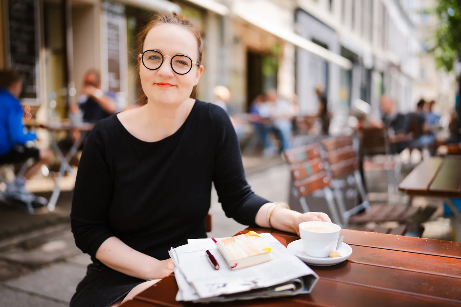 Insa van den Berg, Frau mit roten Haaren und Brille, mit Kaffee und Zeitung in einem Straßencafé.