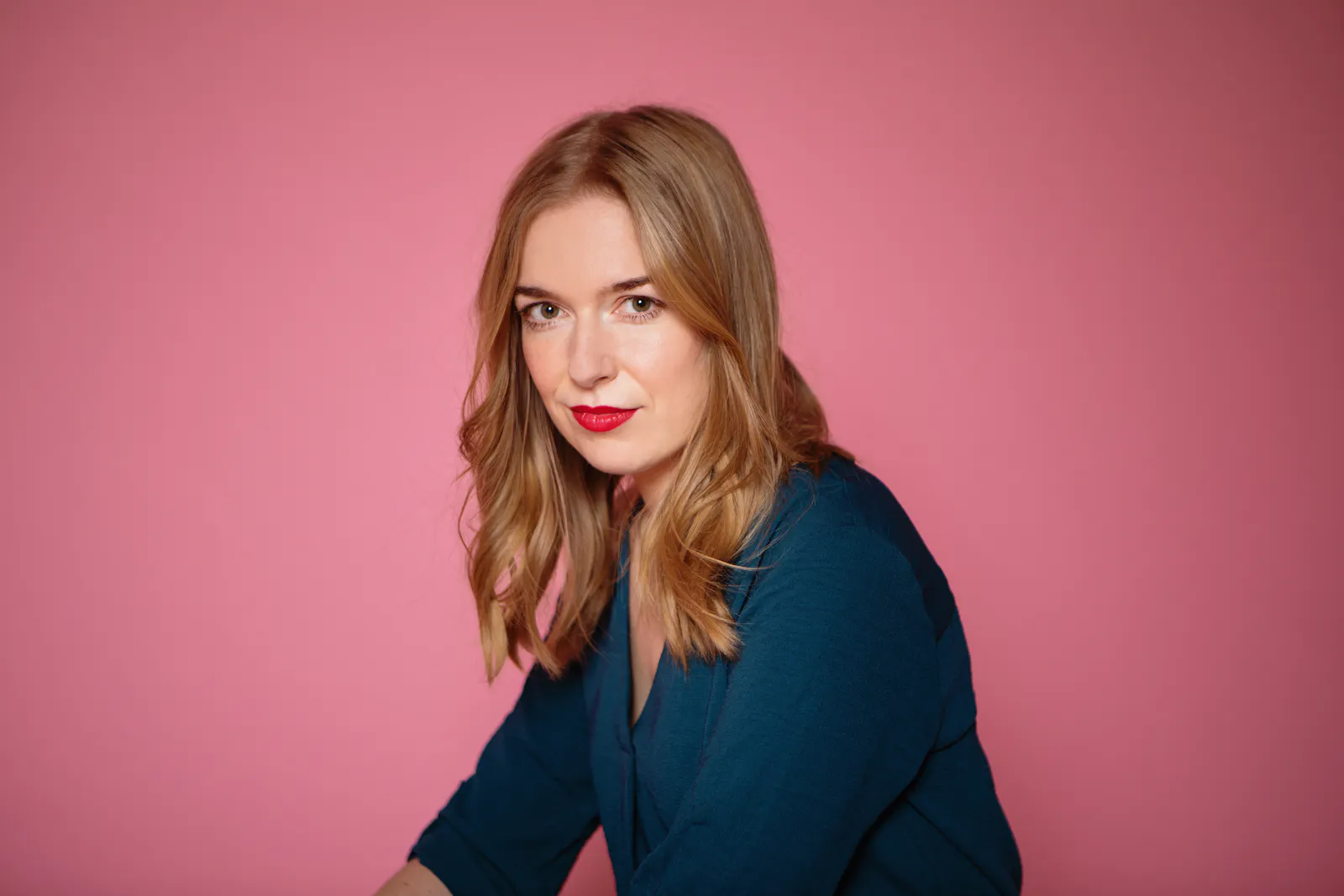 Ein Porträt der Journalistin Anne Dittmann: Sie hat langes, blondes Haar, das sie offen trägt. Sie trägt eine petrolblaue Bluse und roten Lippenstift und schaut selbstbewusst in die Kamera. Der Hintergrund ist pink. (c) Birte Filmer