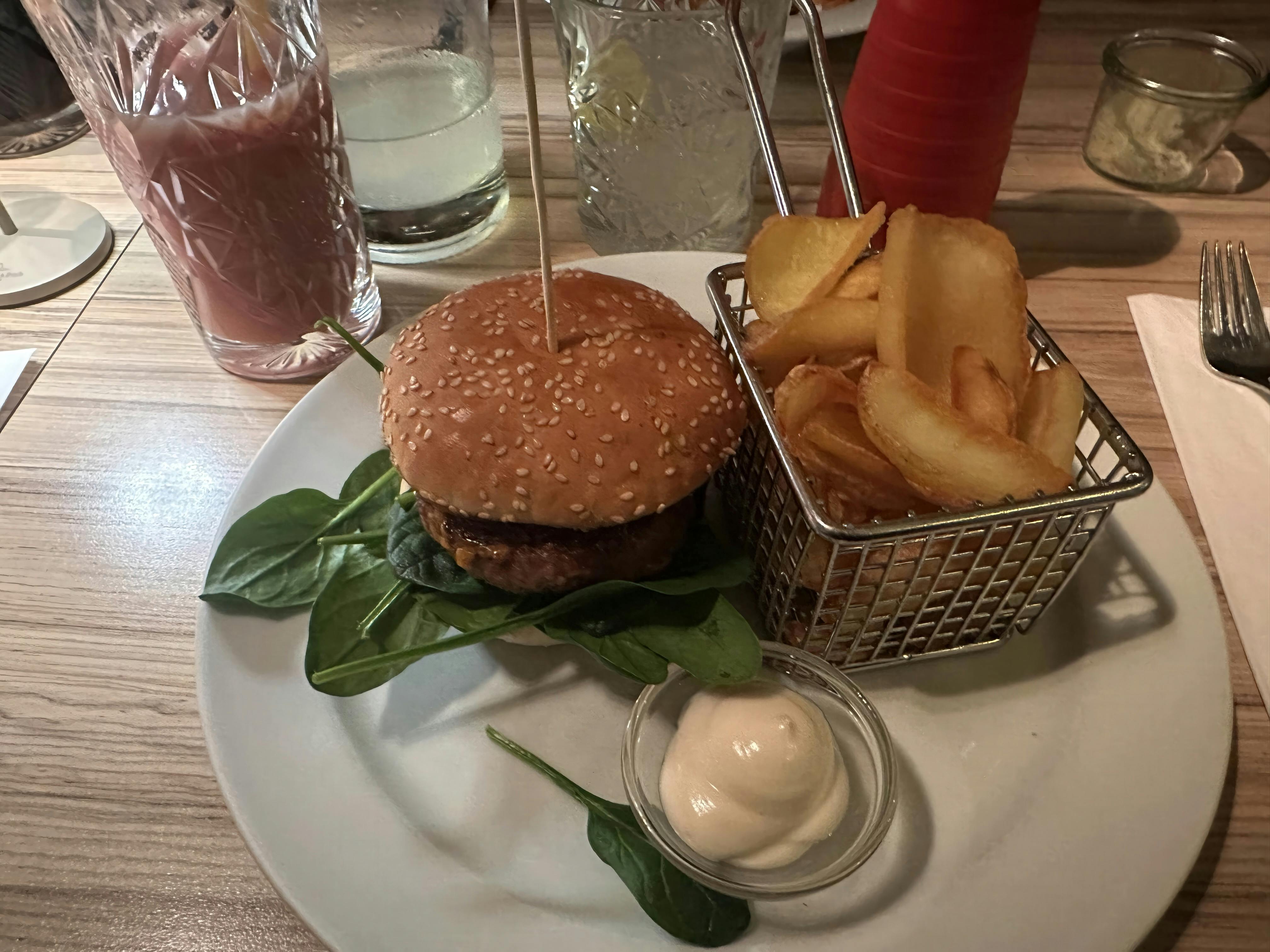 Ein Burger mit Sesambrötchen auf einem Teller, serviert auf frischem Salat, daneben ein Metallkörbchen mit dicken Pommes und ein Schälchen mit Mayo. Im Hintergrund stehen Gläser mit Getränken.
