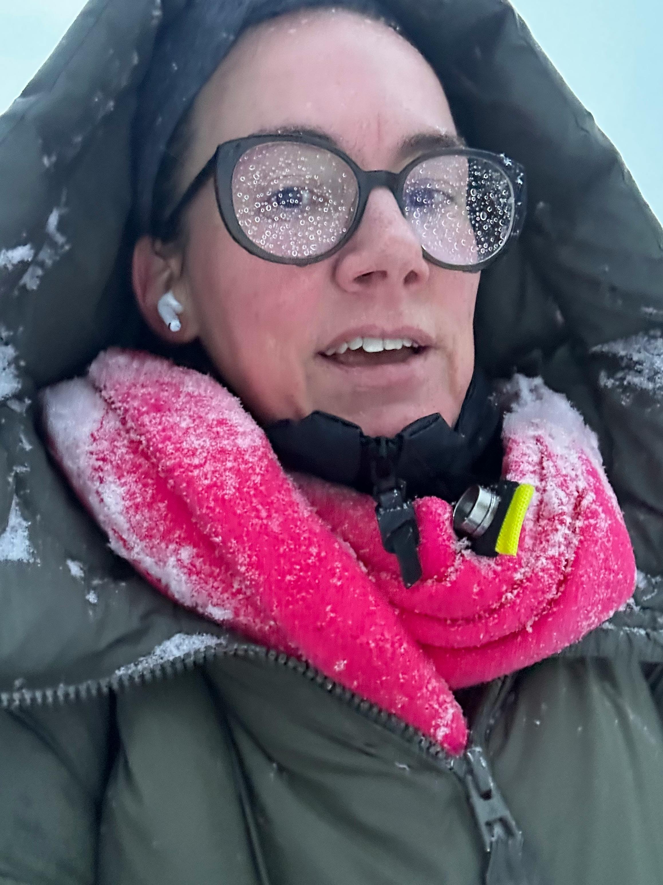 eine Person im grünen Parka, pinkem Schal, Kapuze und Brille ist sehr eingeschneit.