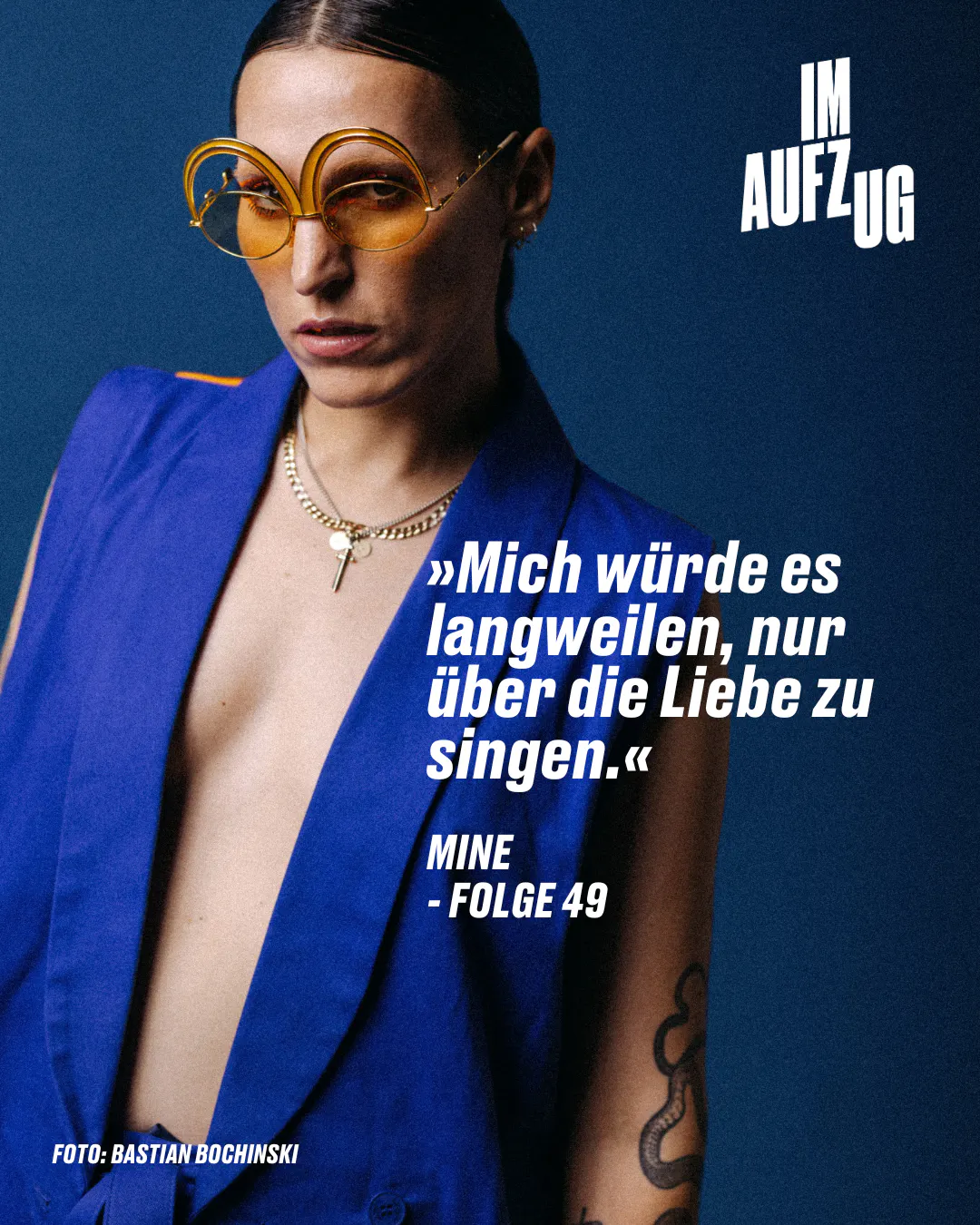 Mine mit einer auffälligen, runden, goldfarbenen Brillen und einem dunkelblauen Blazer. Sie trägt eine Halskette mit einem Anhänger und hat ein sichtbares Tattoo auf ihrem rechten Arm. Ihr Blick ist ernst und direkt in die Kamera gerichtet. Im Hintergrund ist der Text "IM AUFZUG" zu sehen. Unten steht ein Zitat auf Deutsch: "Mich würde es langweilen, nur über die Liebe zu singen."