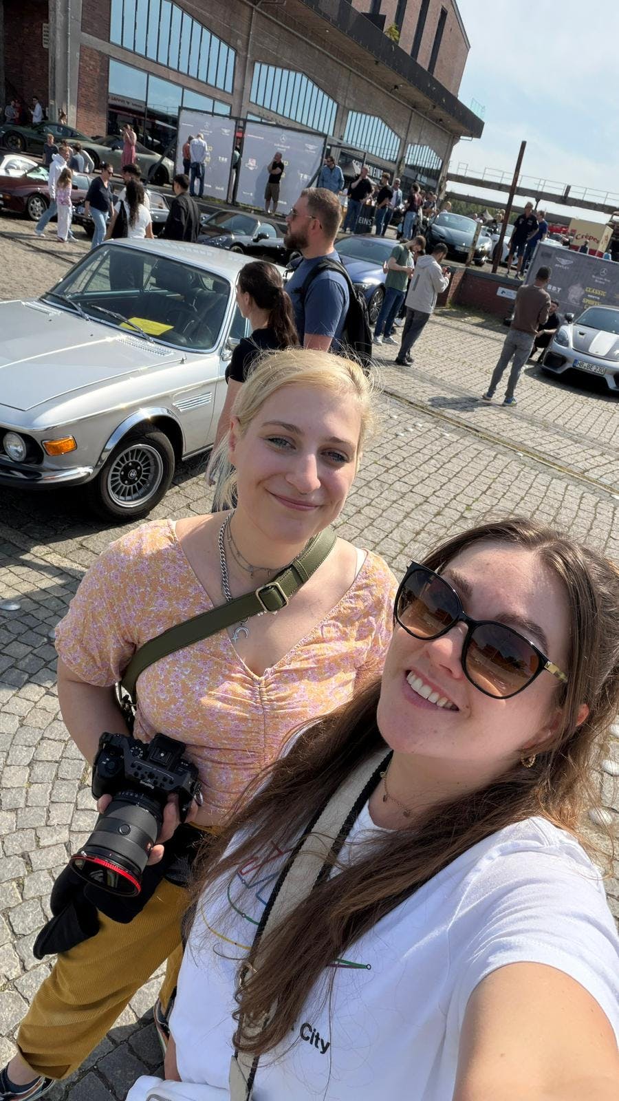 Zwei junge Frauen vor einem silbenren BMW auf einem offenen Platz, im Hintergrund laufen Menschen herum
