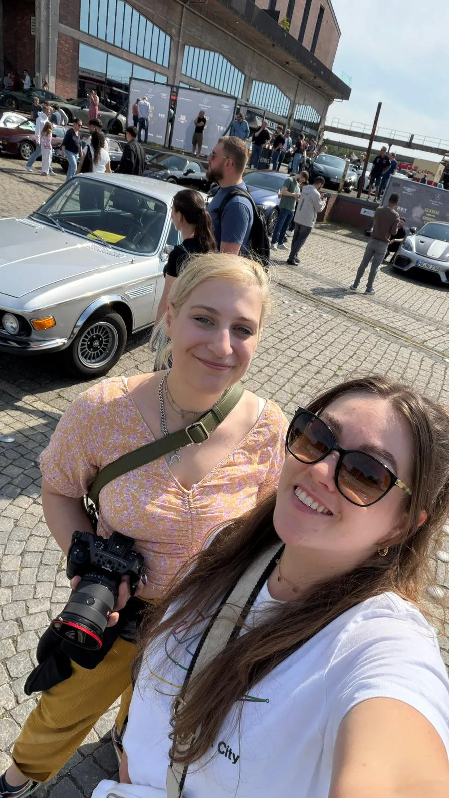 Zwei junge Frauen vor einem silbenren BMW auf einem offenen Platz, im Hintergrund laufen Menschen herum