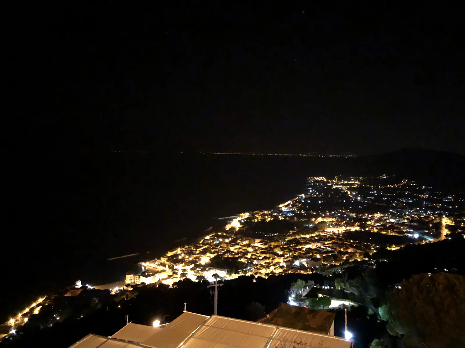 Nächtlicher Blick von einem Hügle auf einen beleuchteten Ort, links davon das nachtschwarze Meer