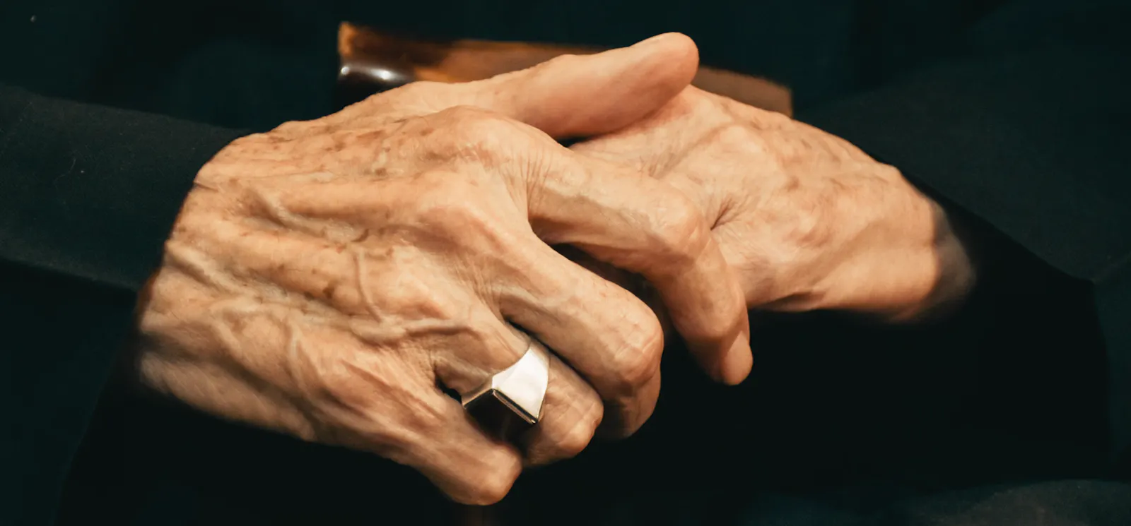 Nahaufnahme von den gefalteten Händen einer älteren Person. Die Hände sind von Altersflecken und Falten gezeichnet und tragen einen auffälligen Silberring. Der Hintergrund ist dunkel und lässt die Hände im Fokus erscheinen