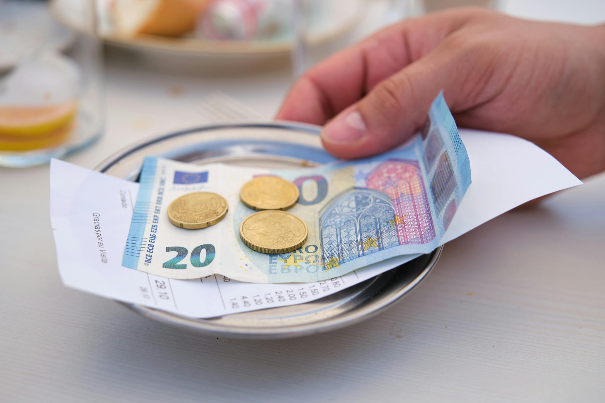 Das Bild zeigt eine Hand, die Bargeld von einem Teller nimmt. Auf dem Teller liegen ein 20-Euro-Schein, einige Münzen und ein Kassenbon. Im Hintergrund sind unscharf Teller mit Essen zu erkennen. Die Szene spielt in einem Restaurant oder Café.