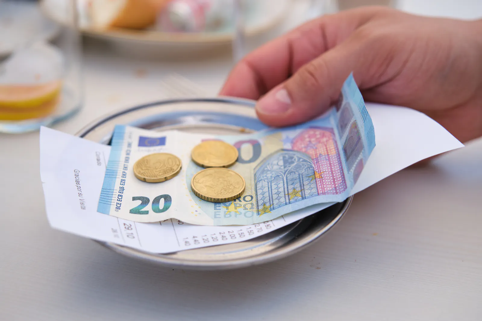 Das Bild zeigt eine Hand, die Bargeld von einem Teller nimmt. Auf dem Teller liegen ein 20-Euro-Schein, einige Münzen und ein Kassenbon. Im Hintergrund sind unscharf Teller mit Essen zu erkennen. Die Szene spielt in einem Restaurant oder Café.