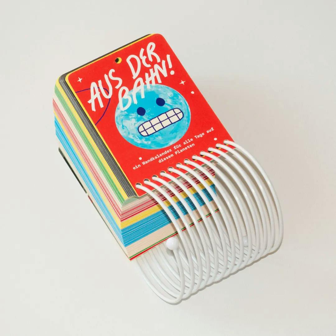 Small spiral-bound flip calendar with a red cover showing a blue planet with a grimacing face and white text
