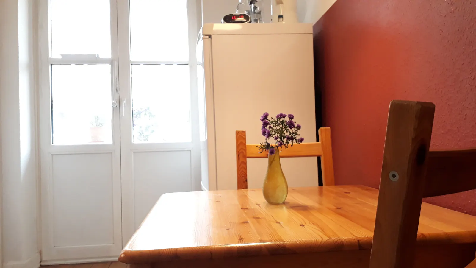 Küchentisch aus hellem Holz mit zwei Stühlen, kleine gelbe Vase mit violetten Blumen darauf. Im Hintergrund Balkontür und Kühlschrank.