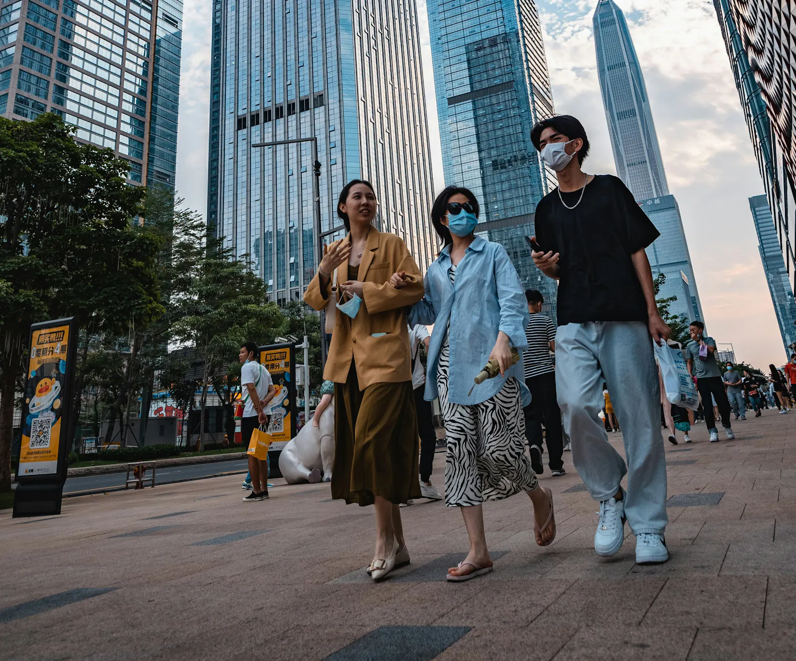 Drei junge Menschen in einer asiatischen Großstadt. Sie tragen Mundmasken und hinter ihnen ragen Hochhäuser auf.