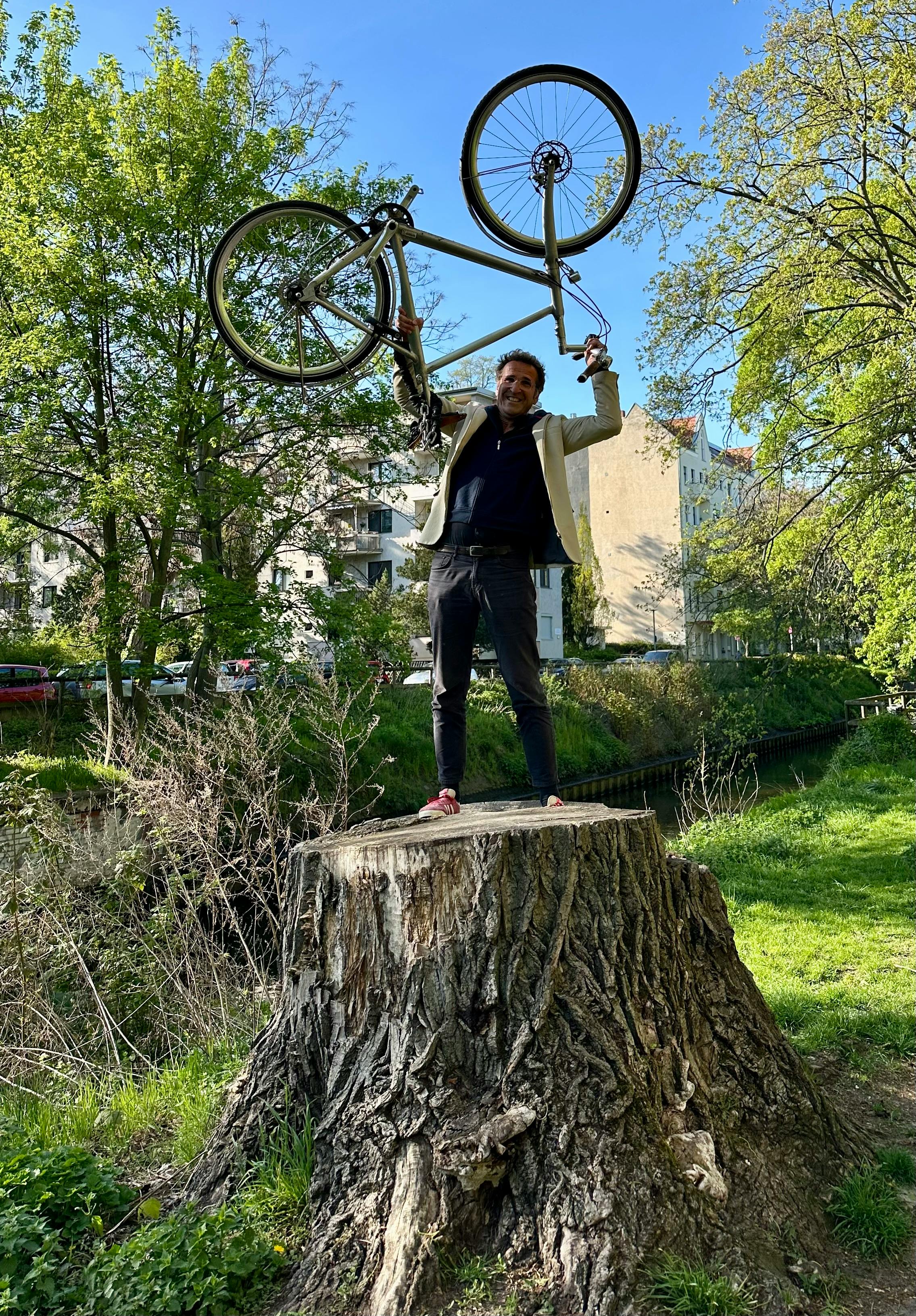 Ein Mann steht auf einem großen Baumstumpf. Er stemmt ein Fahrrad über sich die Höhe.